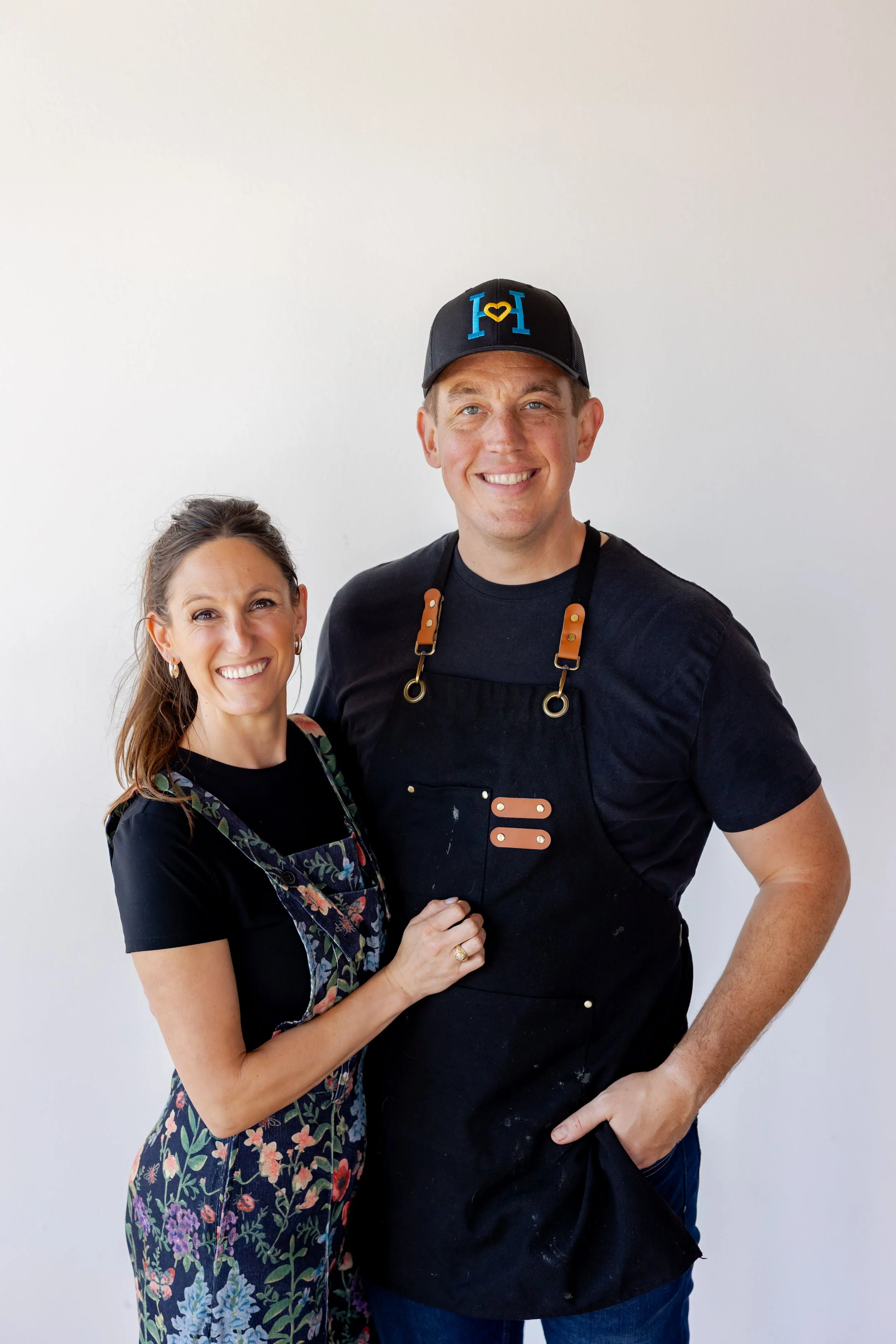 A smiling woman with brown hair in a dress with floral patterns and a black apron standing next to a smiling man wearing a black baseball cap, black T-shirt, and black apron with leather straps, against a plain white background.