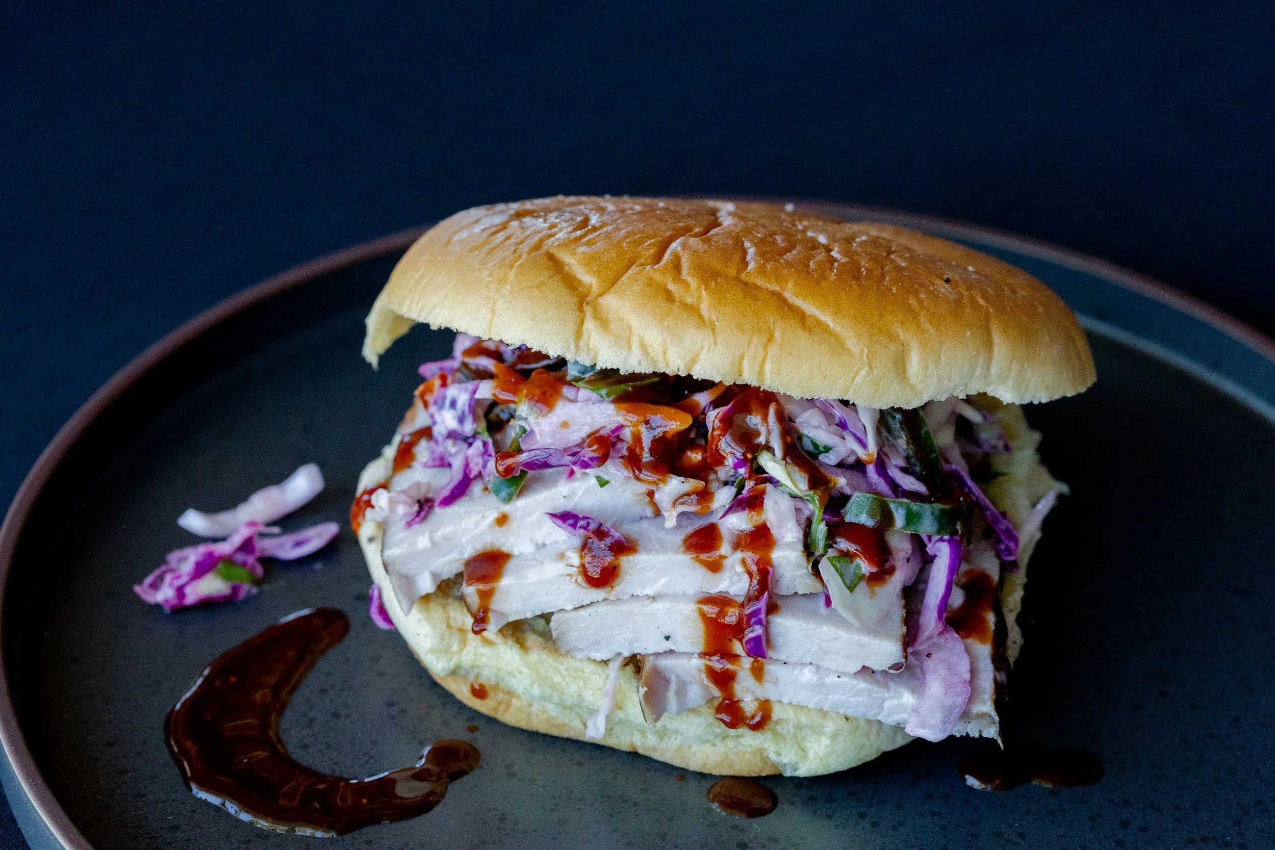 A sandwich with coleslaw, drizzled with sauce, served on a black plate with a dark background.