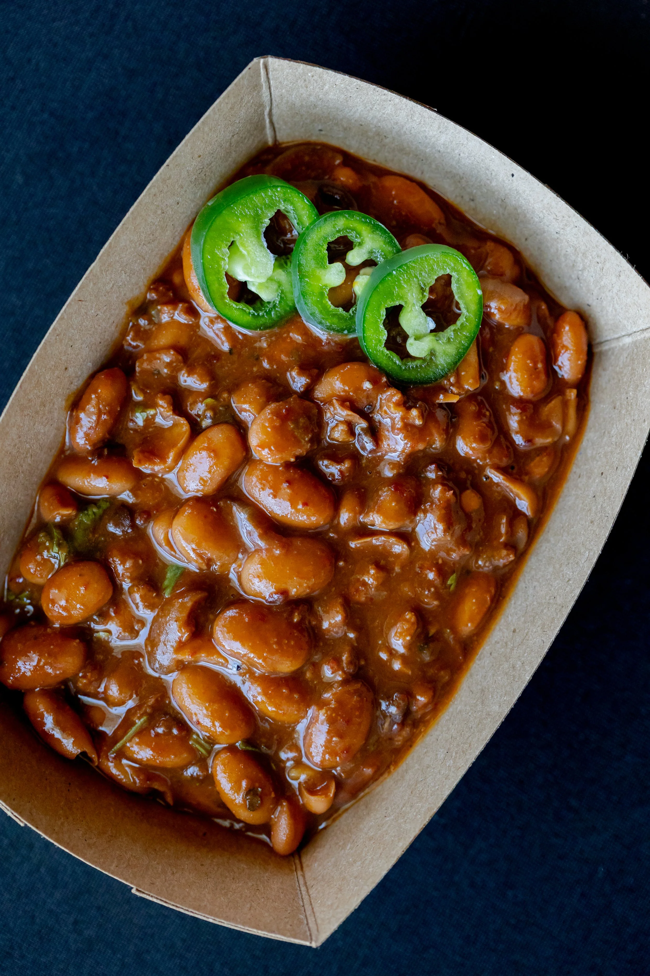 A cardboard food tray containing baked beans in a sauce topped with three slices of green jalapeño peppers.