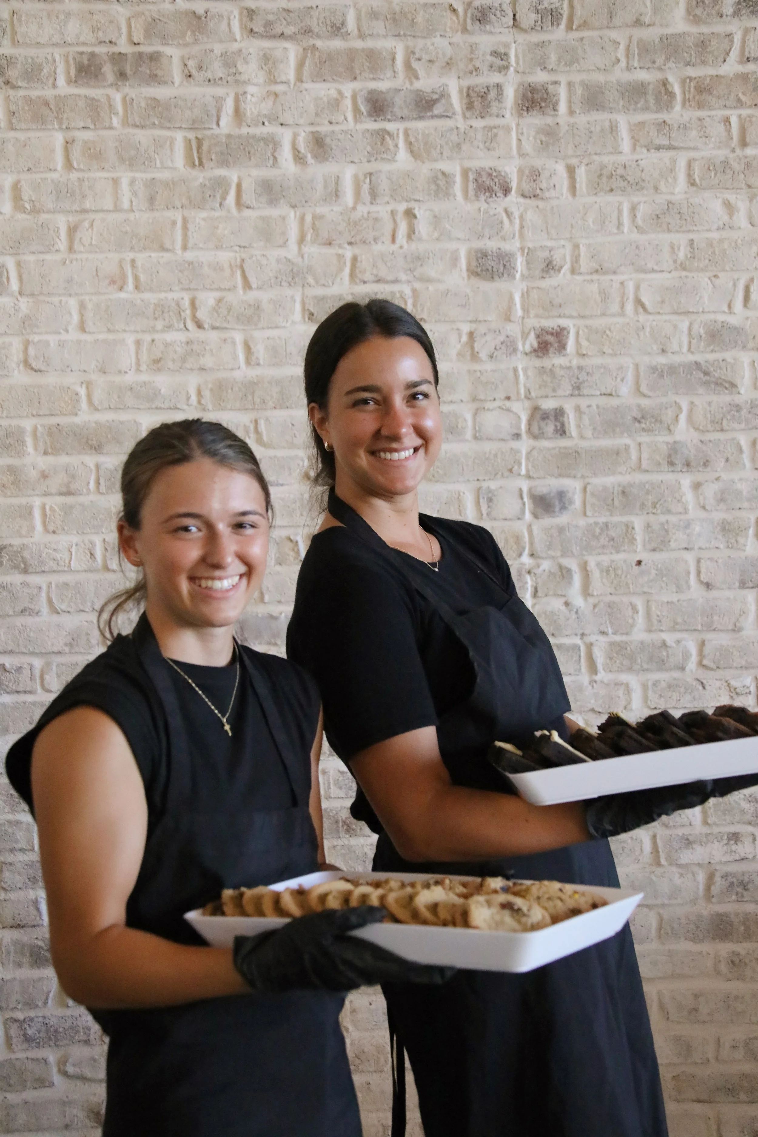 Two women standing in front of a white brick wall, smiling, and holding trays of baked goods, wearing black aprons and gloves.