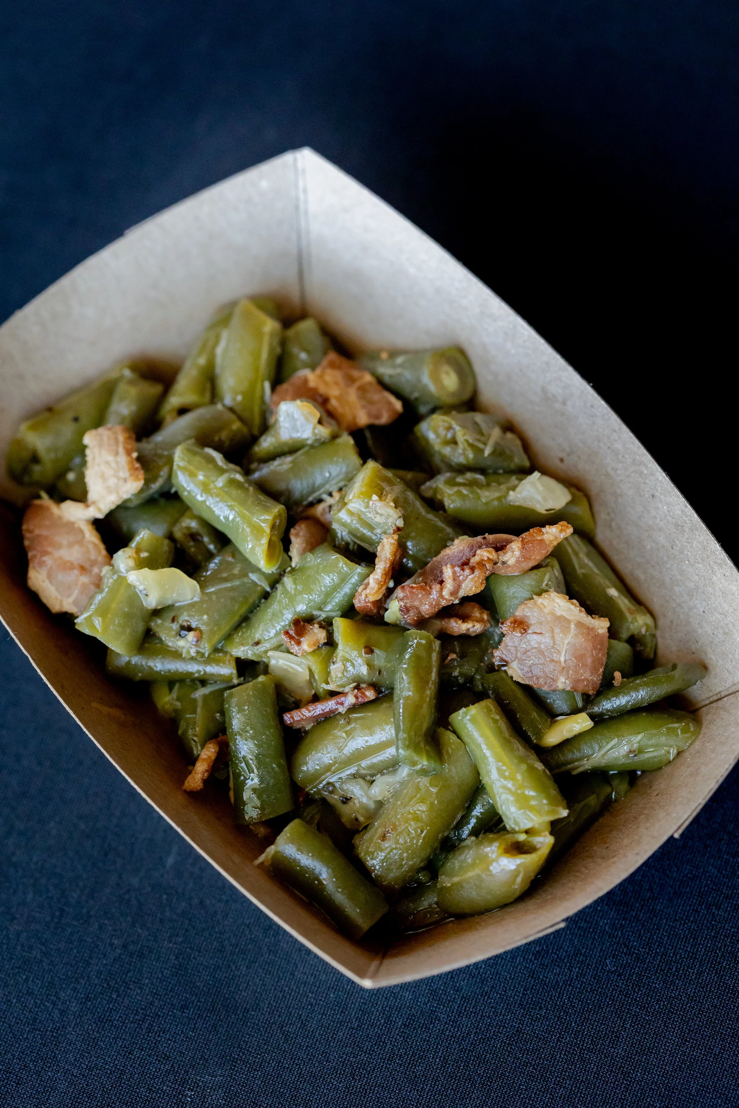 Close-up of cooked green beans with bits of bacon in a paper food tray on a black surface.