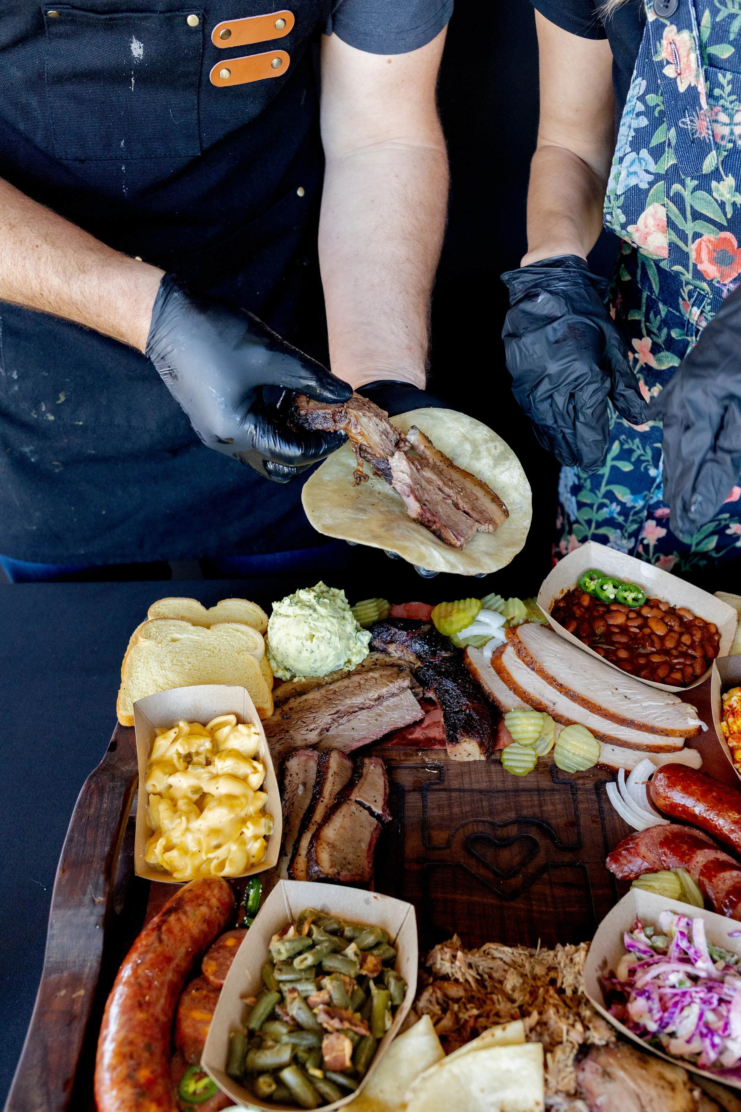 Close-up of a barbecue platter featuring sliced meats, macaroni and cheese, pickles, and various side dishes, with people serving and preparing food in the background.