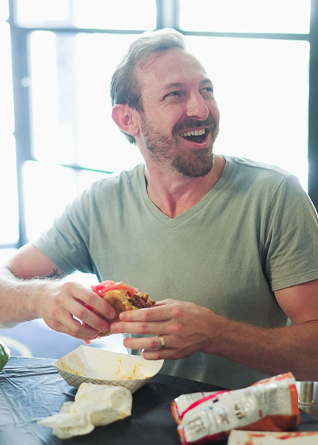 A man with gray hair and a beard, wearing a light gray V-neck T-shirt, sitting at a table, holding a sandwich and laughing, with food and snack wrappers on the table.