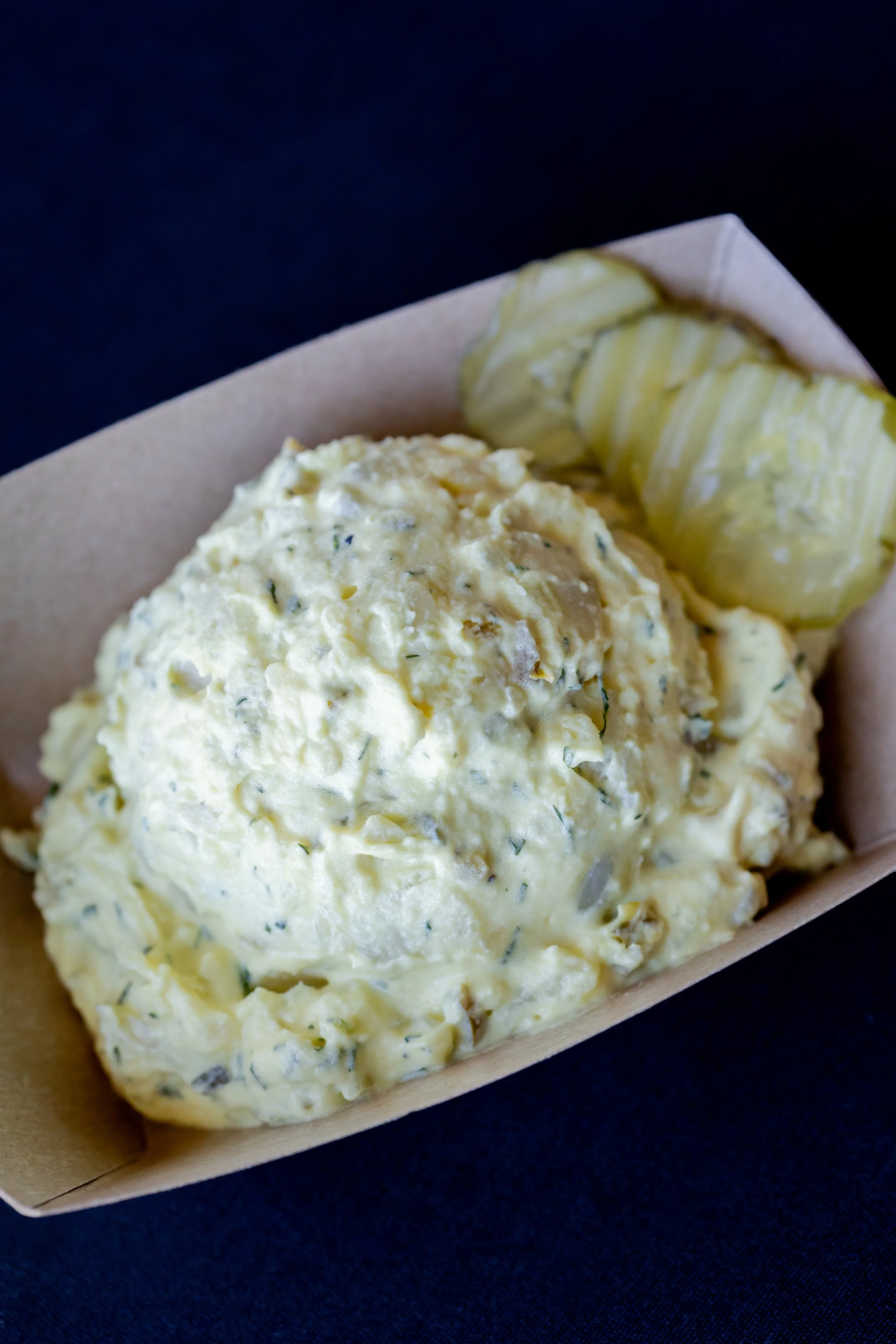 A serving of potato salad with chopped herbs served in a paper tray, with pickle slices on the side.