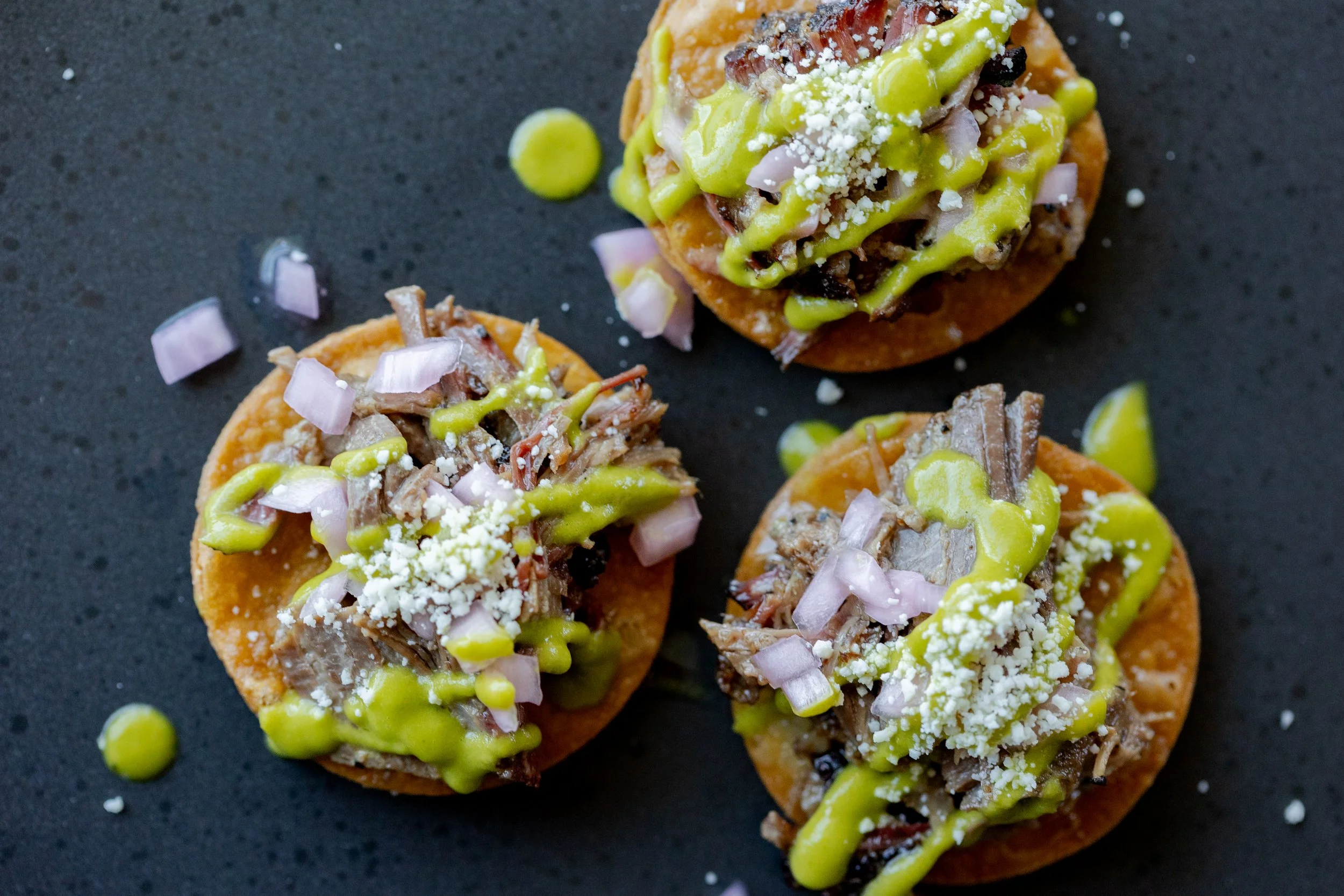 Close-up of shredded meat topped with green sauce, crumbled cheese, and diced onions on small round tostadas, with scattered pieces of onions and green sauce on black surface.