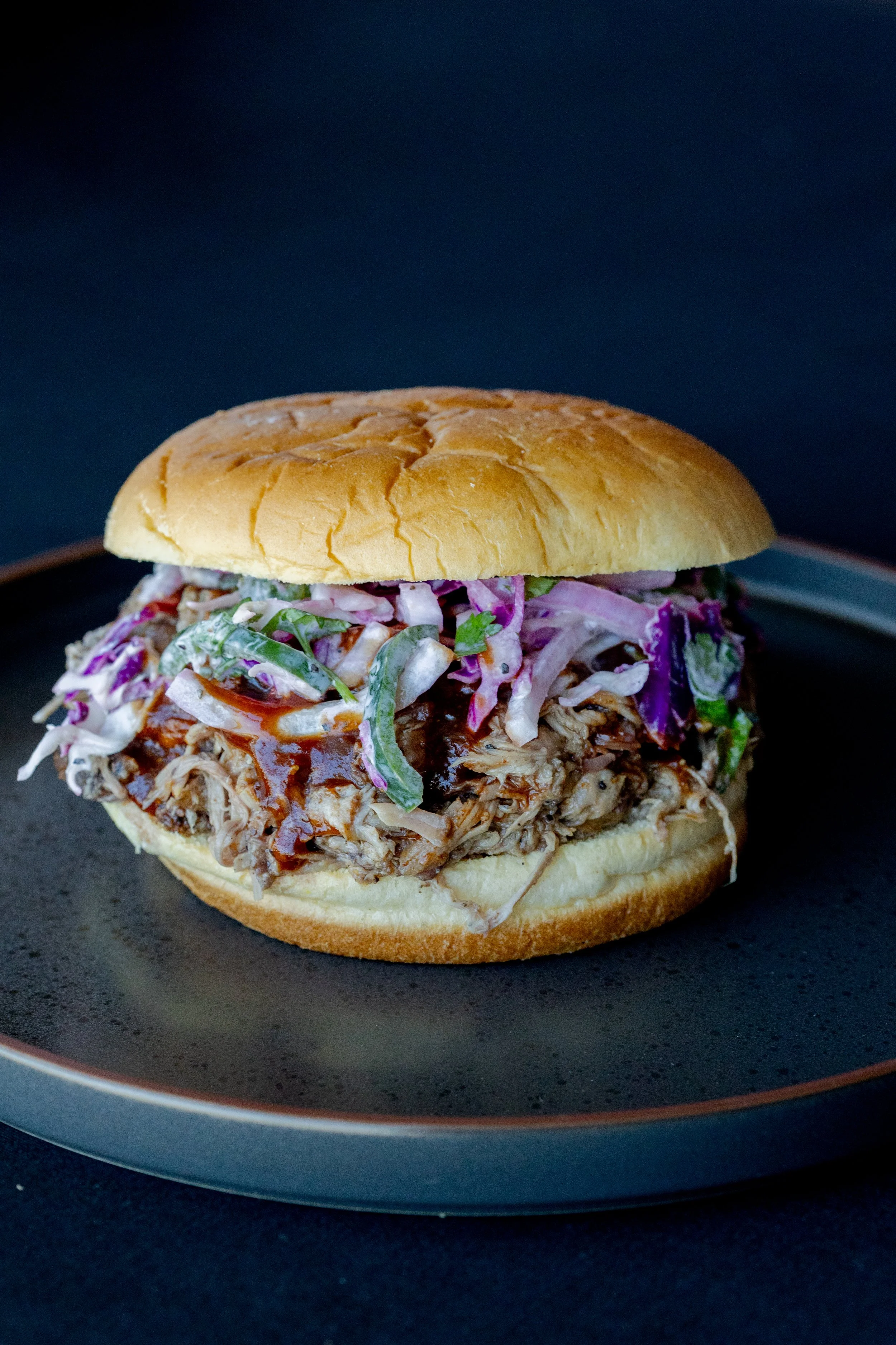 Close-up of a pulled pork sandwich with coleslaw on a bun, served on a black plate.