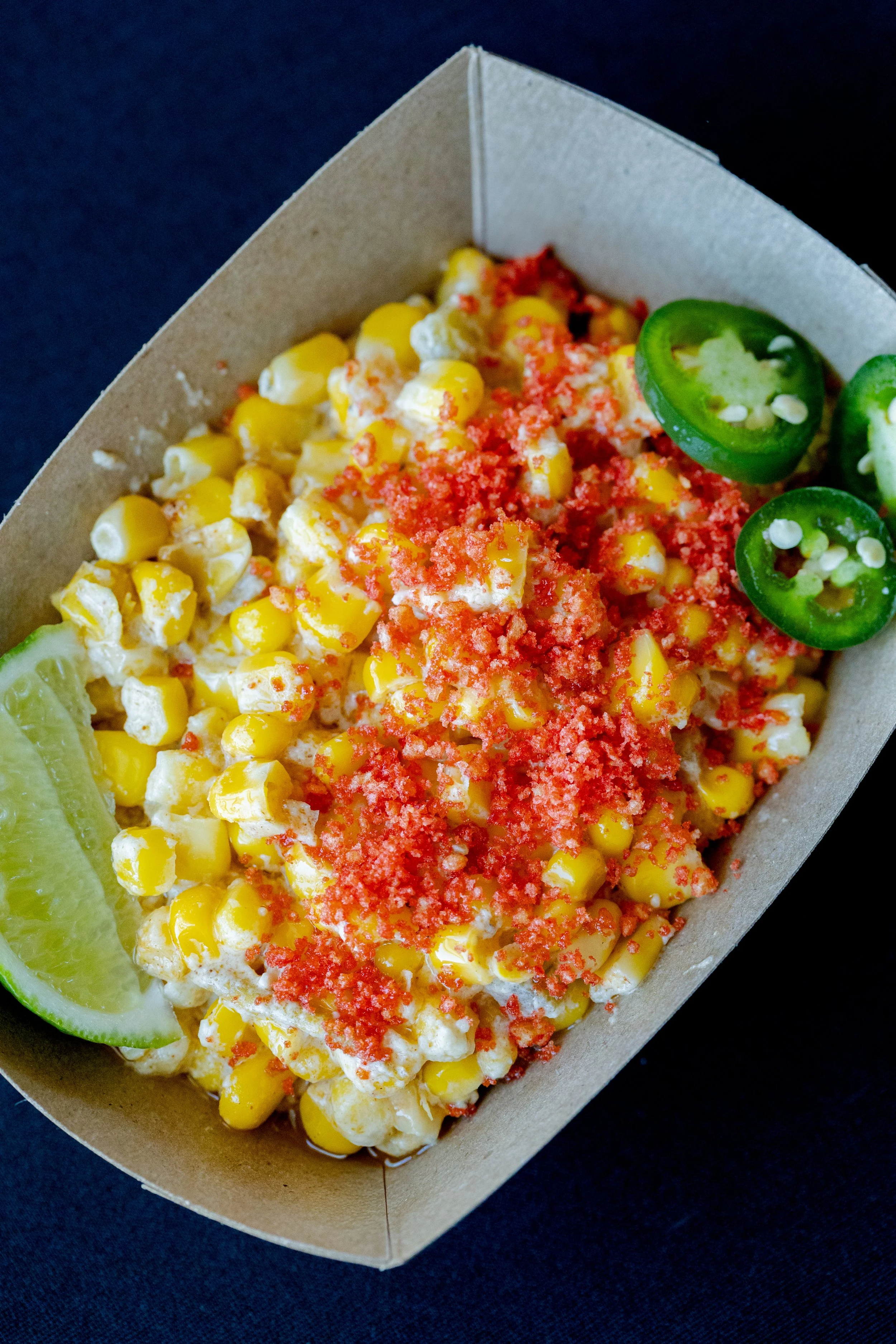 Close-up of a corn salad topped with red seasoning and sliced jalapeños, with a lime wedge on the side in a brown takeout container.