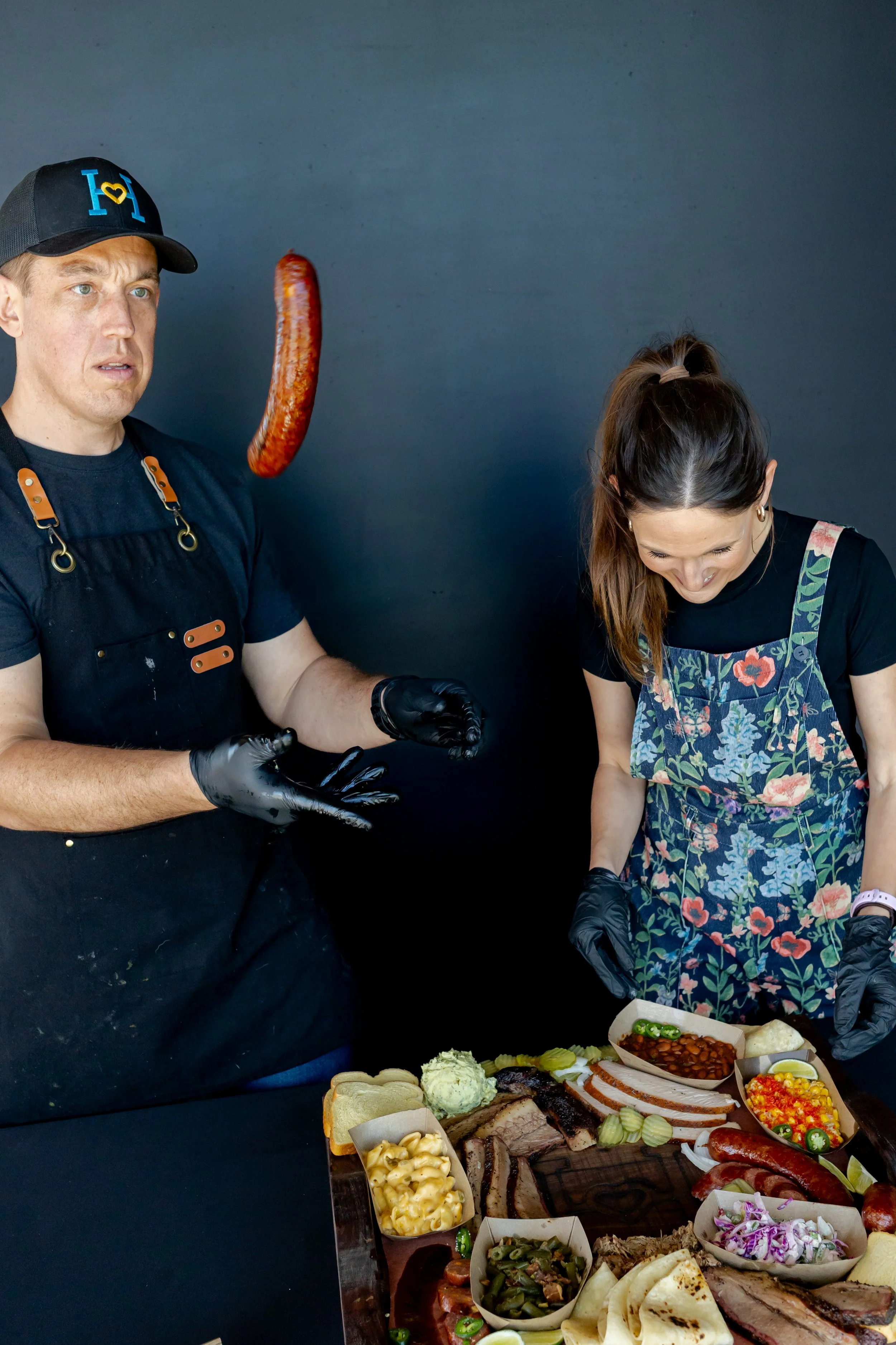 A man and a woman preparing a large platter of various foods, including meats, salads, and side dishes, with a plain navy background.