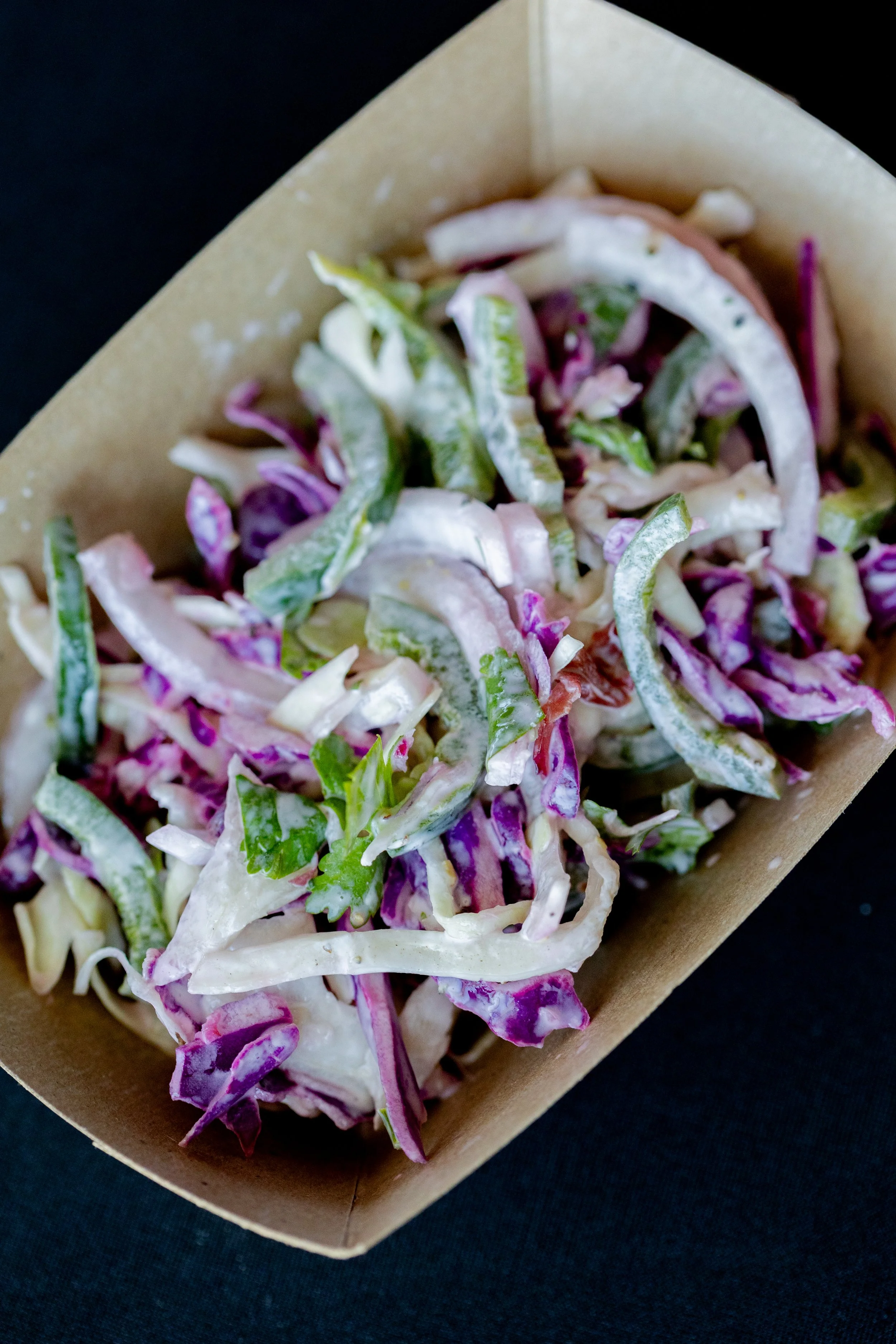 Close-up of a Styrofoam food container with a salad containing shredded purple cabbage, green lettuce, and pieces of white cabbage, placed on a dark surface.