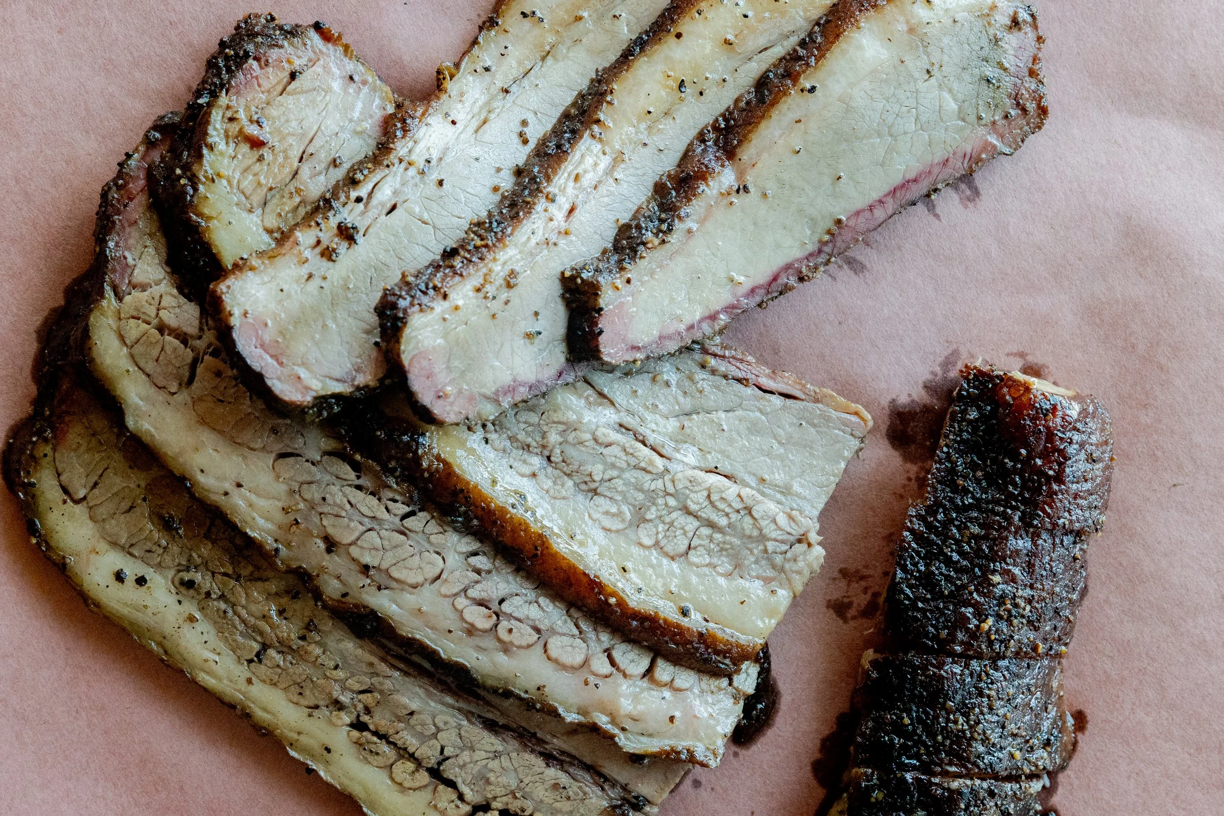 Sliced roasted beef brisket with a dark crust on the outside, resting on pink paper.