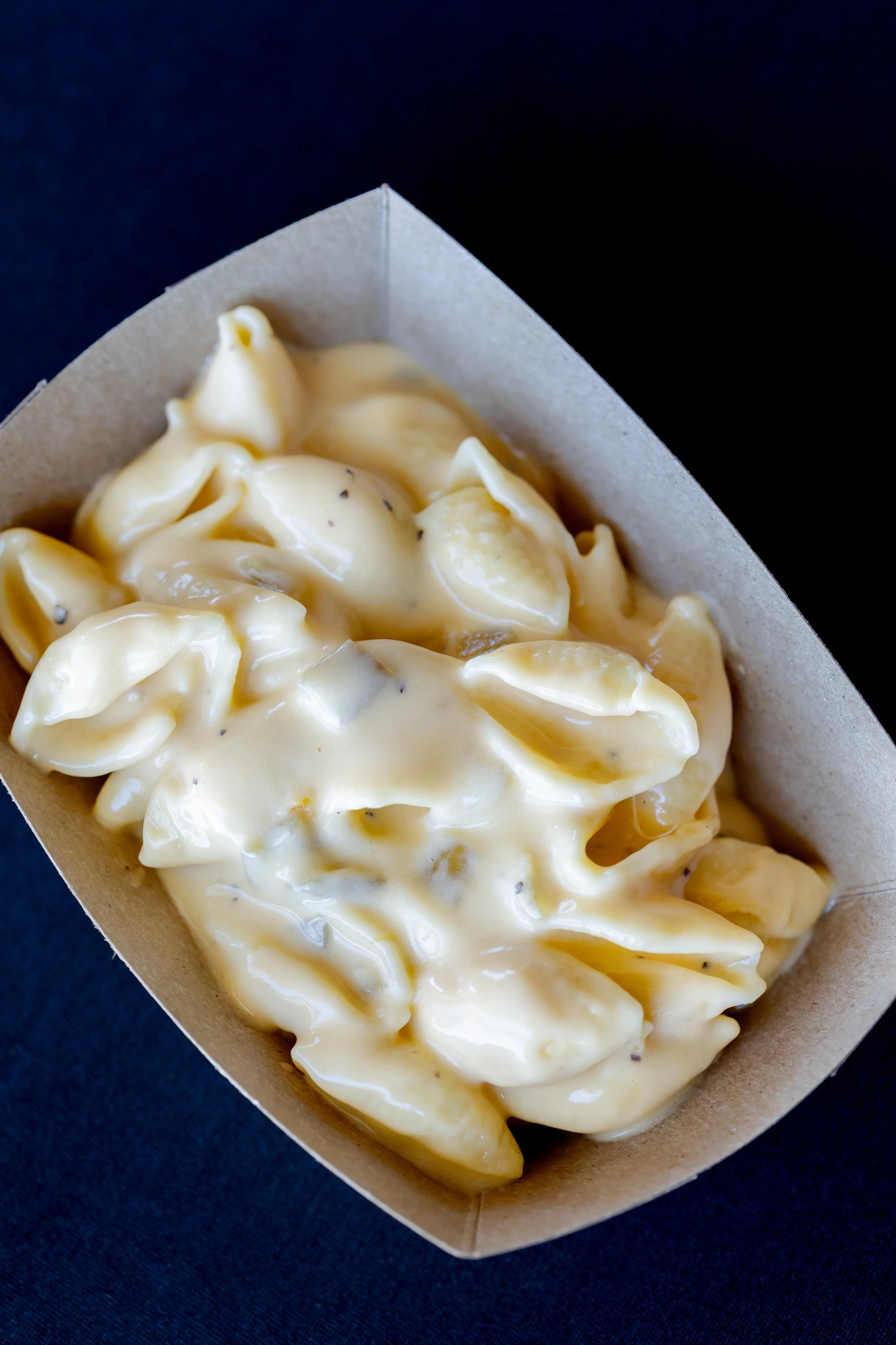 Close-up of a cardboard container holding creamy, yellow cheese tortellini pasta coated in cheese sauce, against a dark background.
