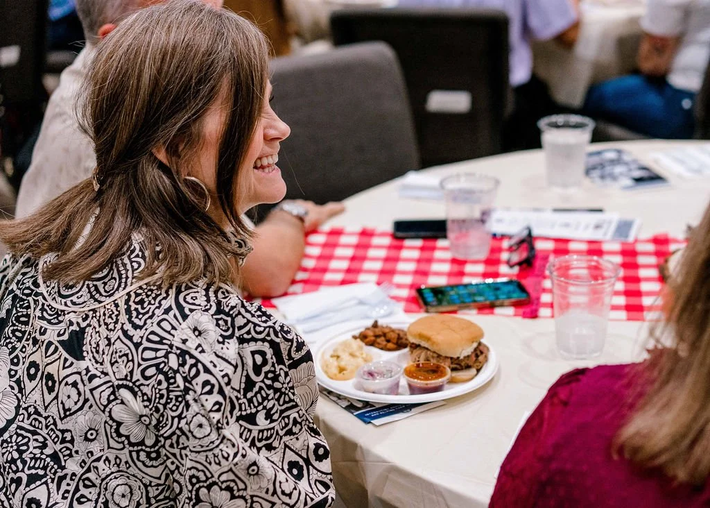 Woman with long brown hair smiling at a woman with blonde hair, sitting at a table with a red and white checkered tablecloth, food, and drinks in a social setting.