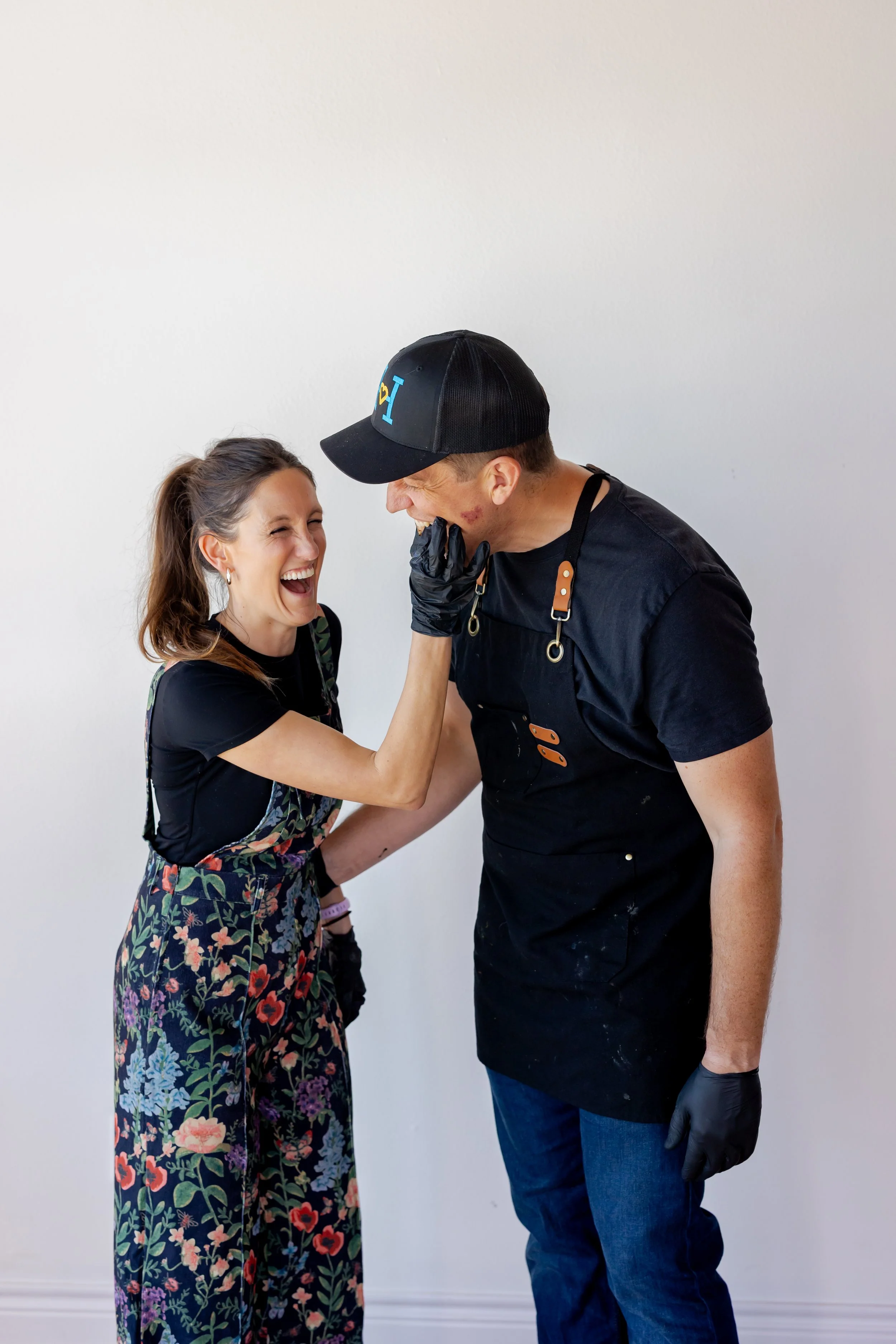 A woman and a man laughing and smiling while hugging each other. The woman has long brown hair and is wearing a black top with a floral dress, while the man is wearing a black cap, black gloves, and a black outfit. They are standing against a white wall.