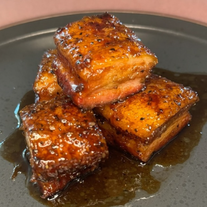 Four glazed pieces of pork belly on a dark plate with sauce.
