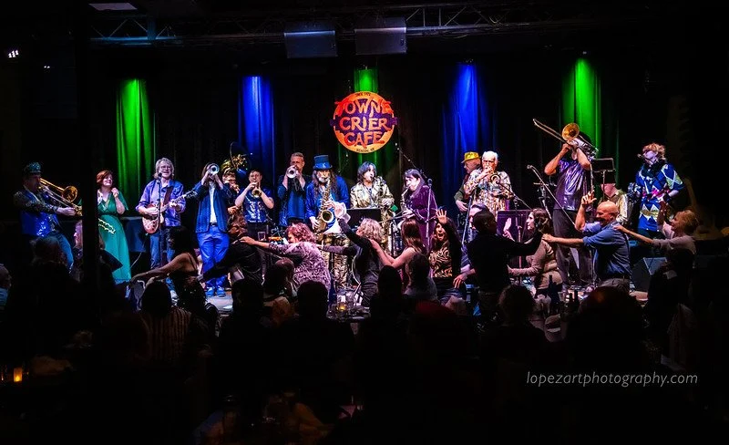 A live musical performance at the Towner Crier Cafe featuring a band with musicians playing brass and woodwind instruments on stage, with an audience dancing and enjoying the show.