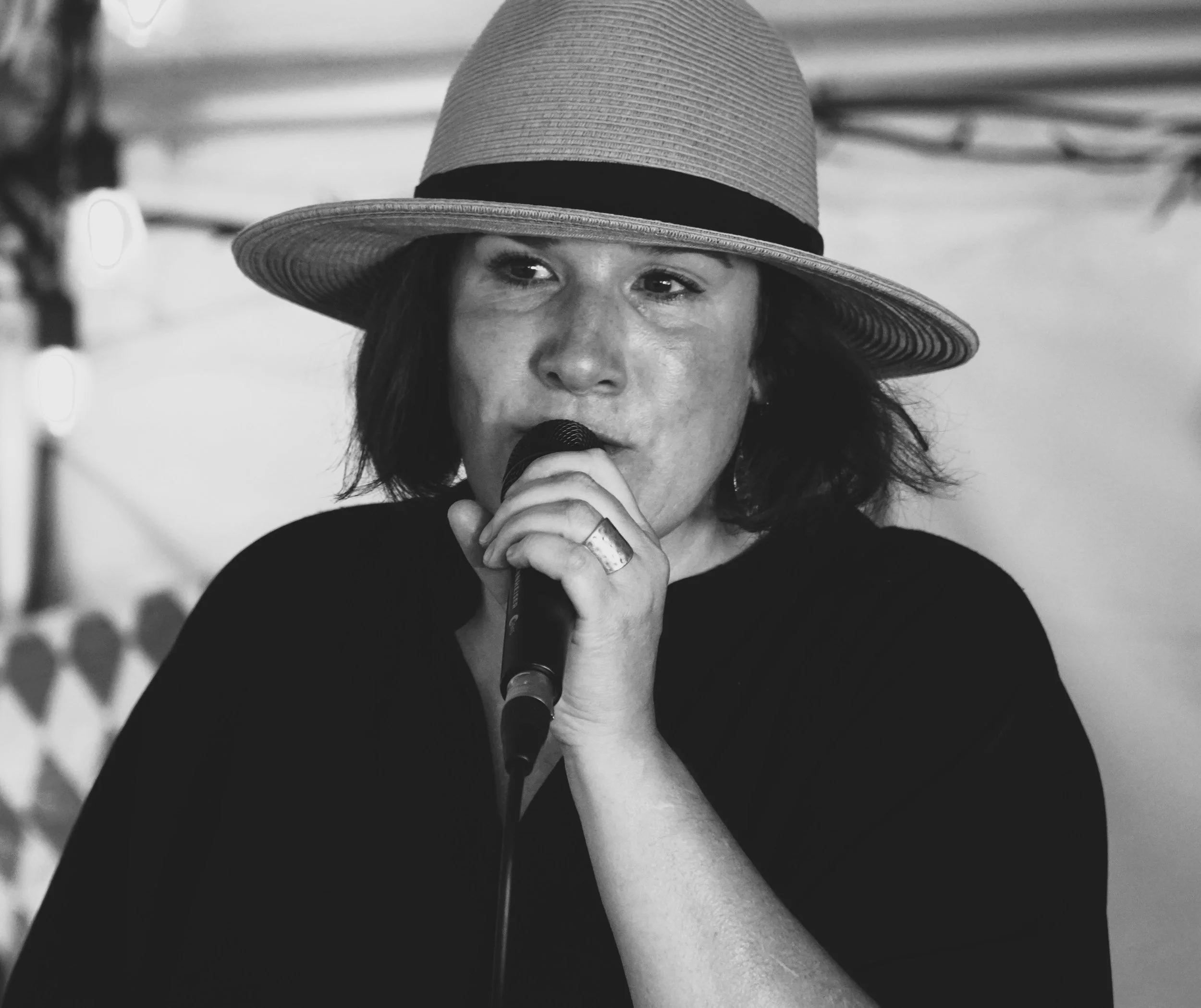 A woman singing into a microphone, wearing a wide-brimmed hat, with a blurred background.