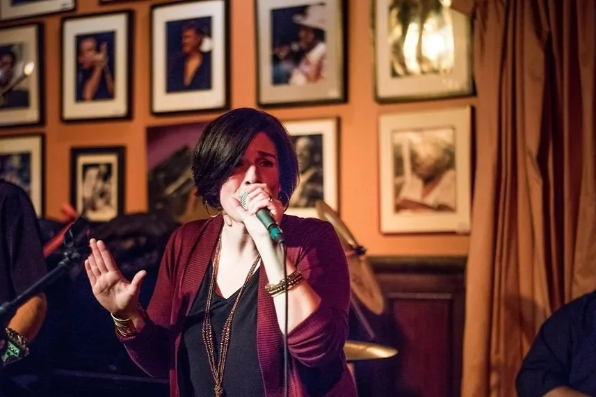 Woman singing into a microphone in a dimly lit venue with framed photos on the wall in the background.
