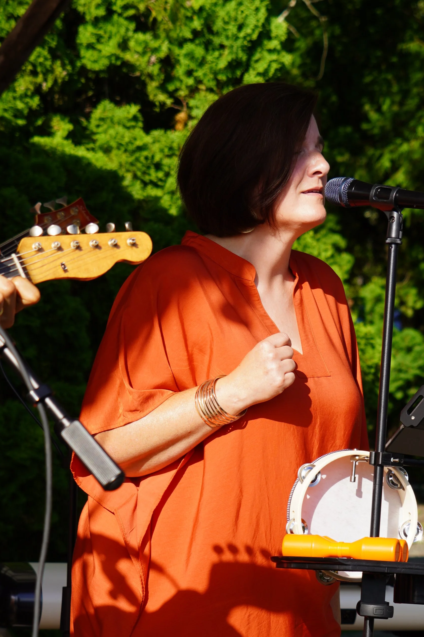 A woman singing into a microphone outdoors, wearing an orange top, with musical instruments including a tambourine and guitar nearby, and lush green trees in the background.