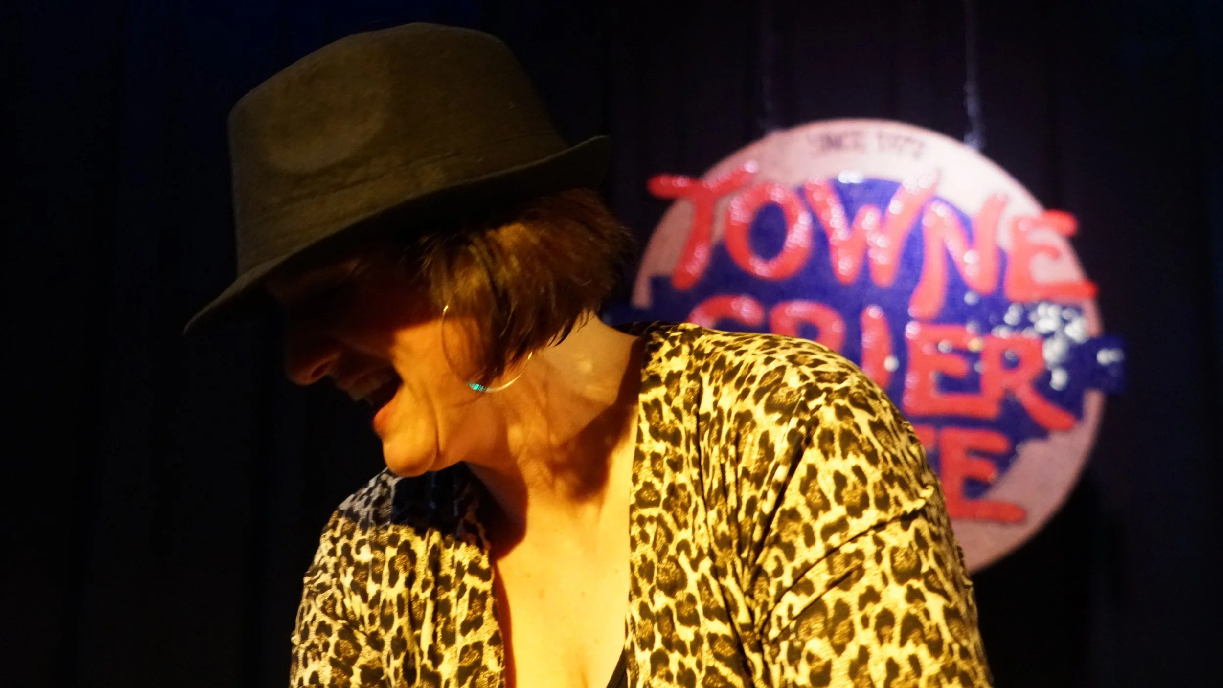 Woman with short dark hair wearing a leopard print top and a dark fedora hat, smiling and looking downward, with a sign in the background that says "TOWN AND COUNTRY" in red and blue letters.