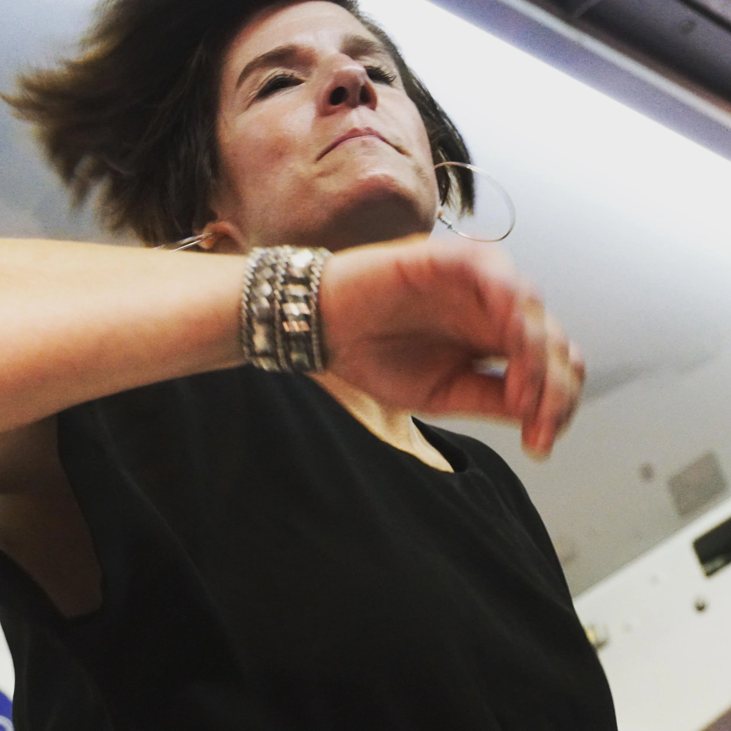 Close-up of a woman with short dark hair wearing large hoop earrings and a silver bracelet, wearing a black top, looking upwards with her arm raised.