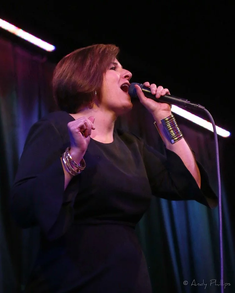 A woman singing into a microphone on stage with her eyes closed and one fist clenched.