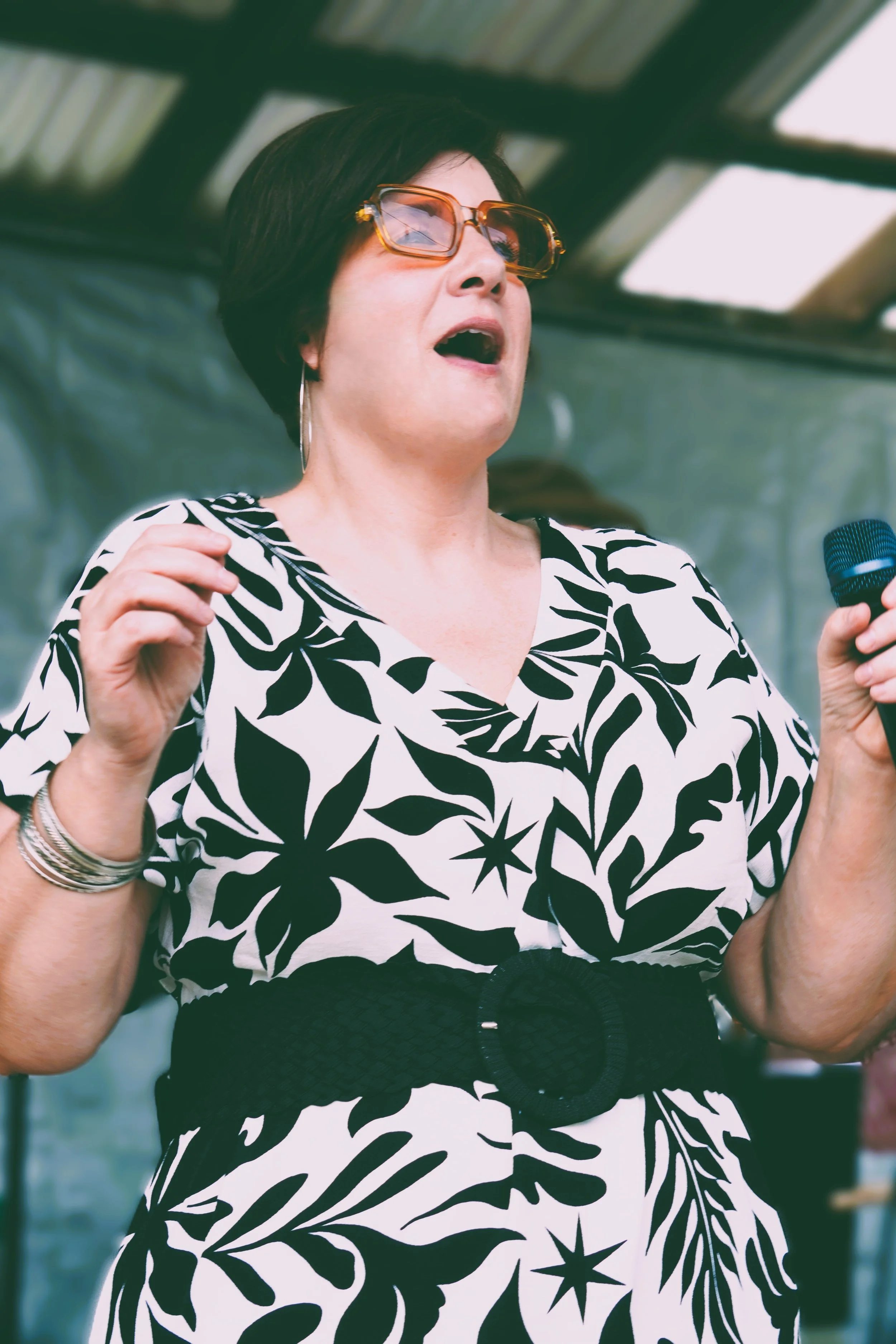 A woman singing into a microphone at an outdoor event, wearing sunglasses, a black and white patterned dress, and a wide belt.
