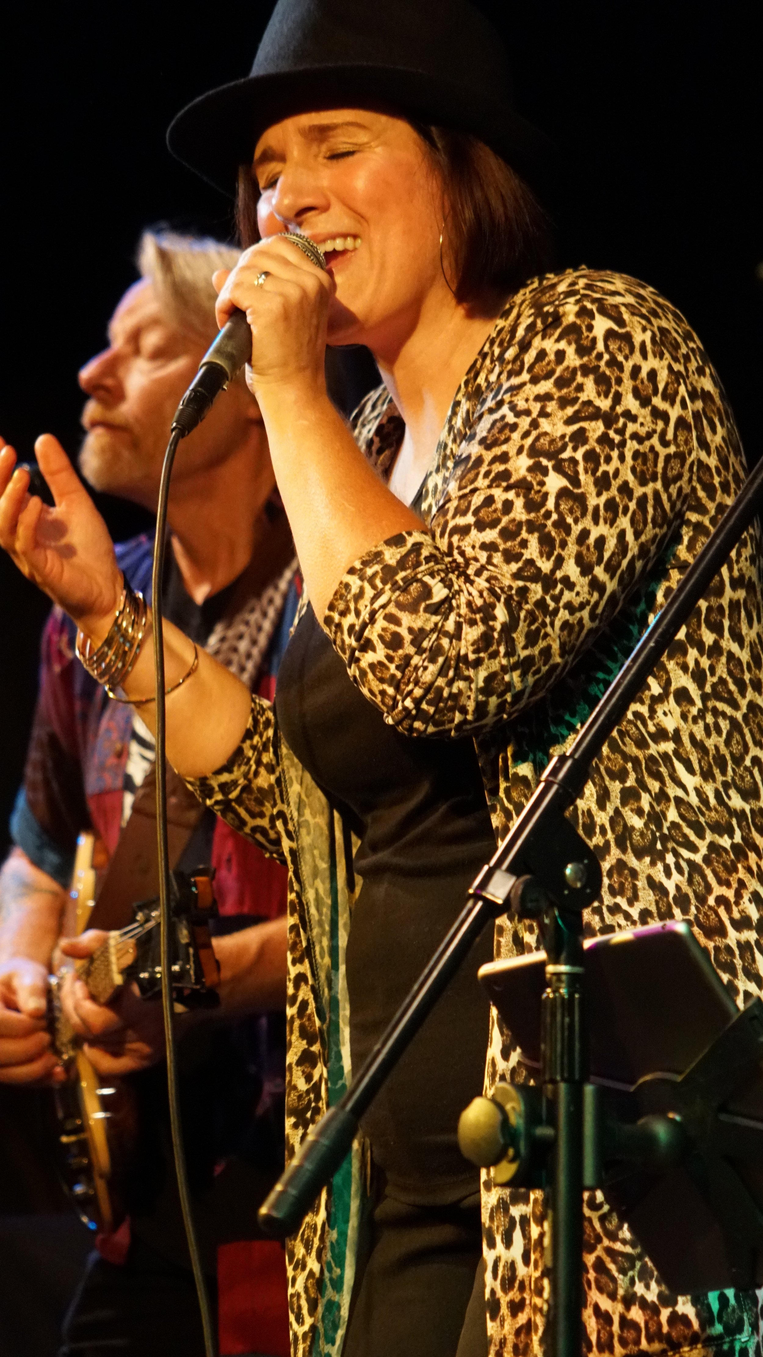 A woman singing into a microphone, wearing a leopard print jacket and a black hat, with a guitarist in the background.