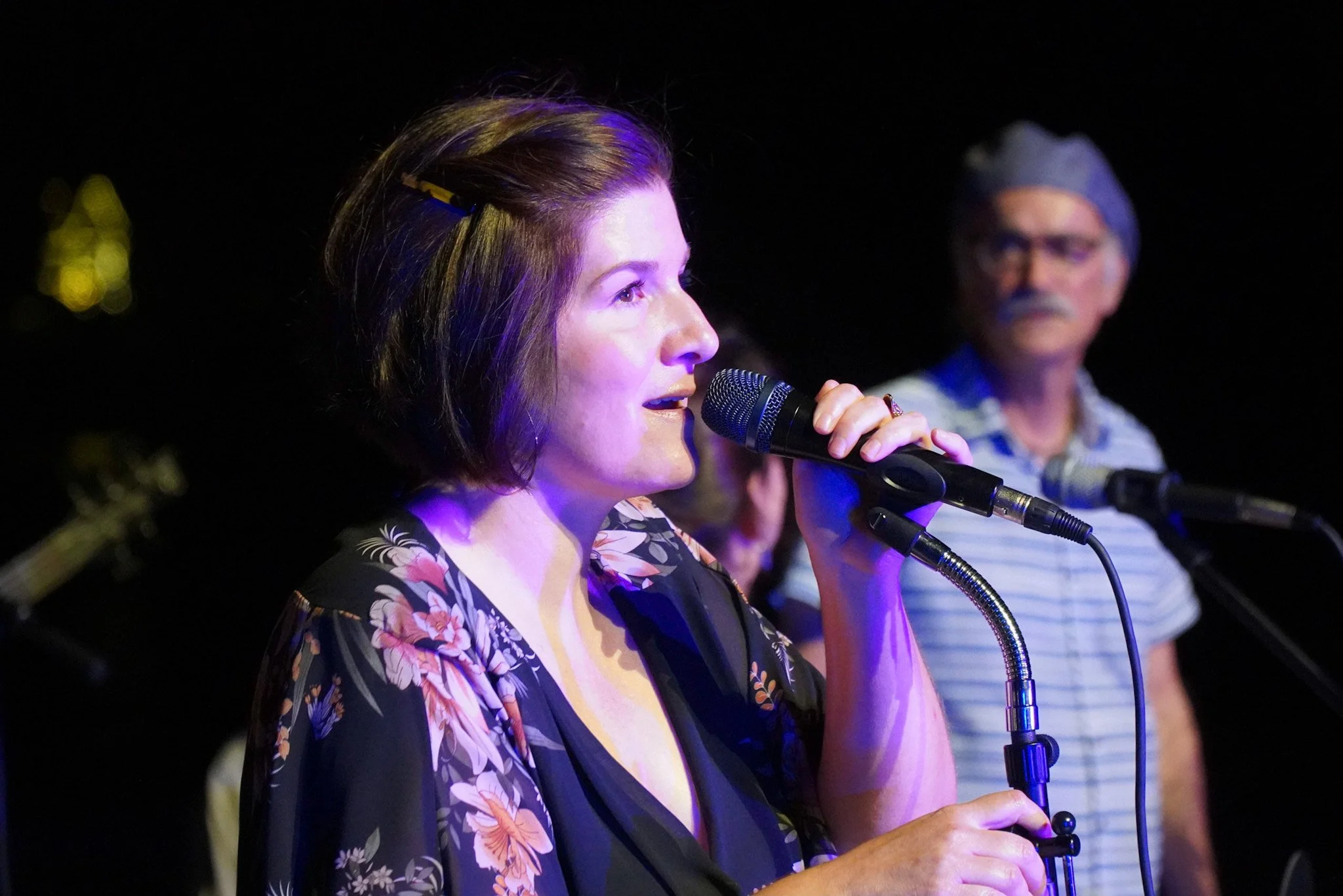 Woman with short brown hair singing into a microphone on stage, wearing a black floral top, with a man in a striped shirt and glasses in the background.