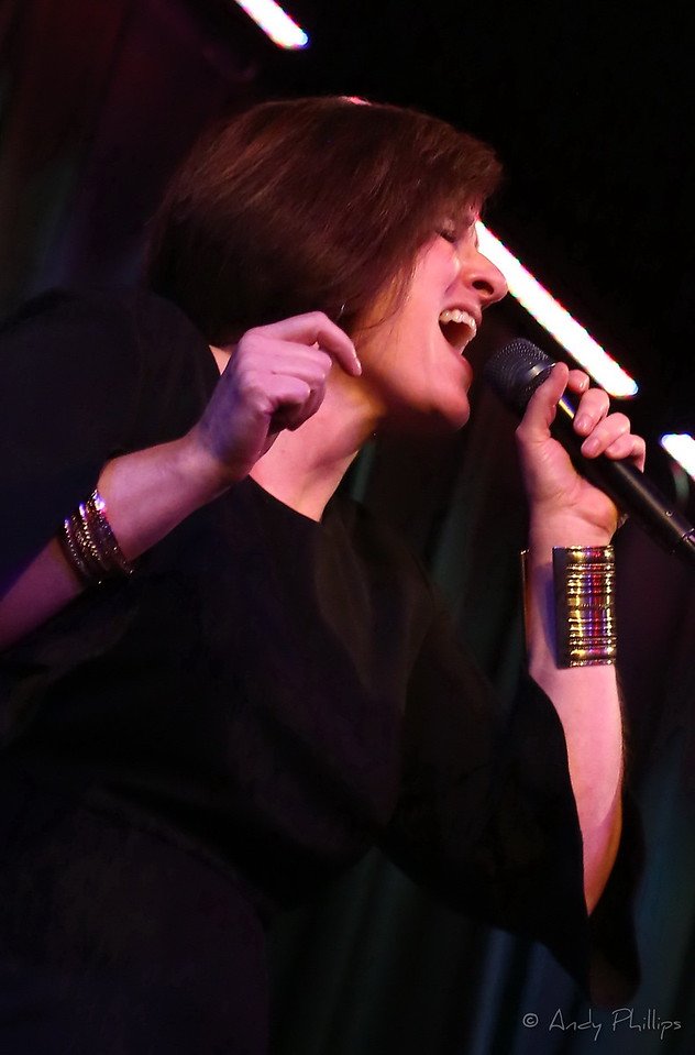 A woman singing passionately into a microphone on stage, with her eyes closed and one fist raised, wearing a black outfit and gold jewelry.