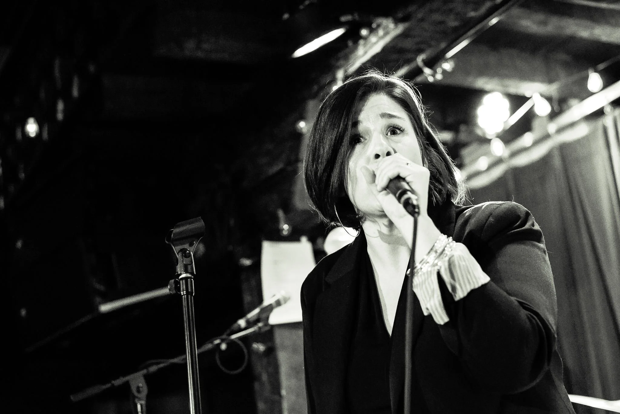 A woman singing into a microphone on stage at a live music venue, with a dark background and stage lighting.