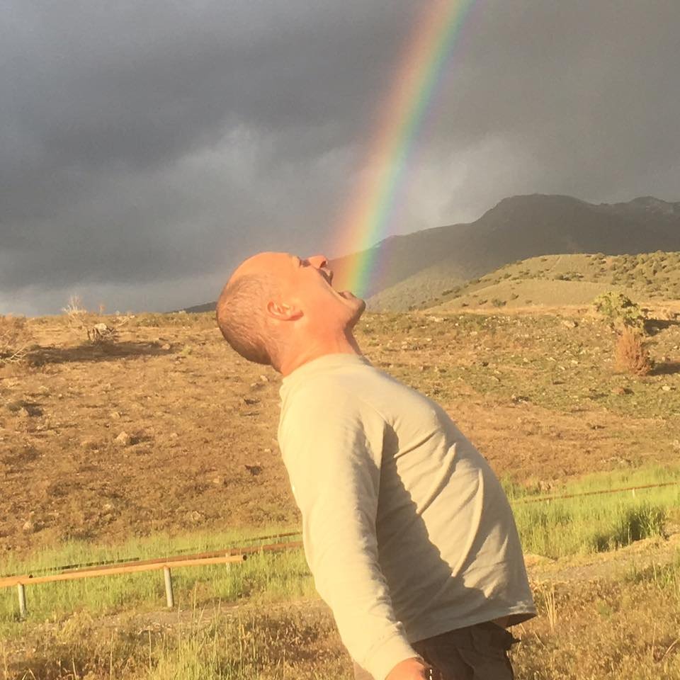 A man standing outdoors with his head tilted back, mouth open as if shouting or laughing, under a rainbow in a cloudy sky, with mountains in the background.