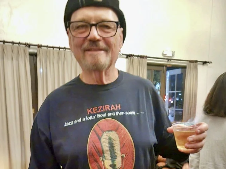 An older man with glasses, a beard, and a mustache wearing a black hat and a dark t-shirt with a graphic of a microphone and text that reads 'KEZIRAH Jazz and a lotta Soul and then some...' holding a small glass of beverage, standing in a room with light-colored curtains and a window in the background.