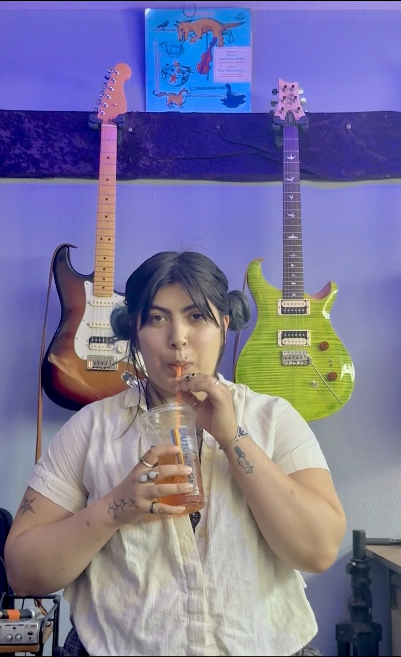 A woman with dark hair in pigtails drinks from a clear plastic cup with a straw, standing in front of two guitars mounted on a wall, one sunburst and one green, with a blue poster featuring dinosaurs above them.