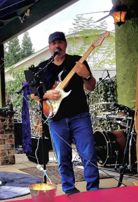 A man wearing a black cap and black shirt playing an electric guitar and singing into a microphone on an outdoor patio surrounded by musical equipment.