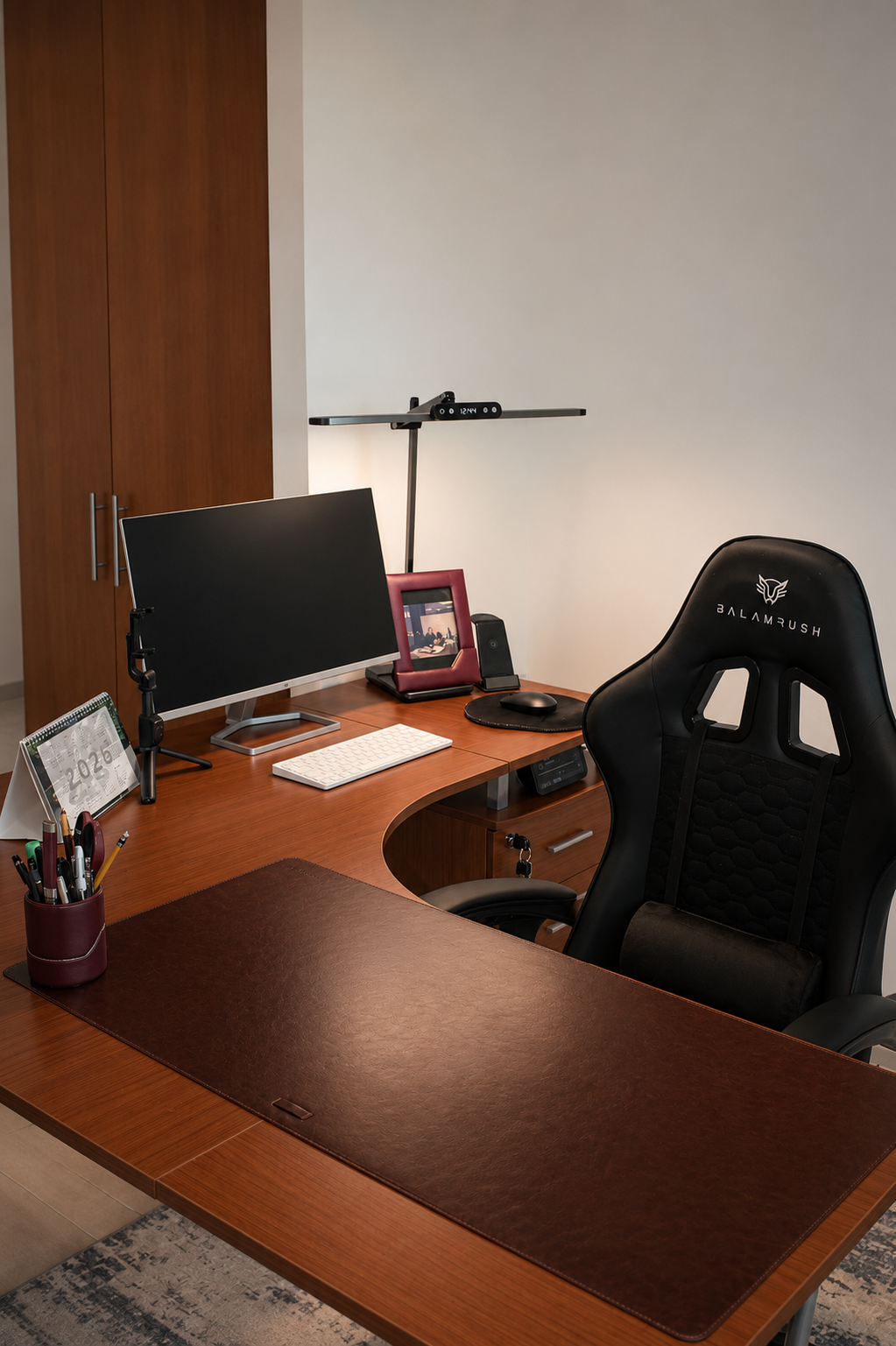 Oficina con escritorio de madera, silla ergonómica negra, monitor, teclado, ratón, lámpara de escritorio, marco de fotos, calendario, y objetos de oficina en un ambiente ordenado y moderno.