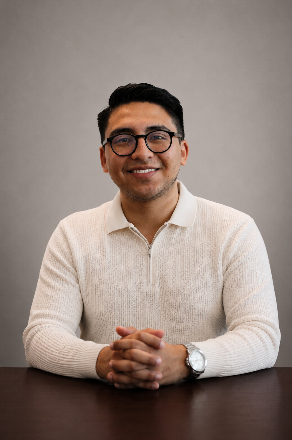 Hombre sonriendo con gafas, usando suéter blanco y reloj, sentado en una mesa frente a una pared gris.