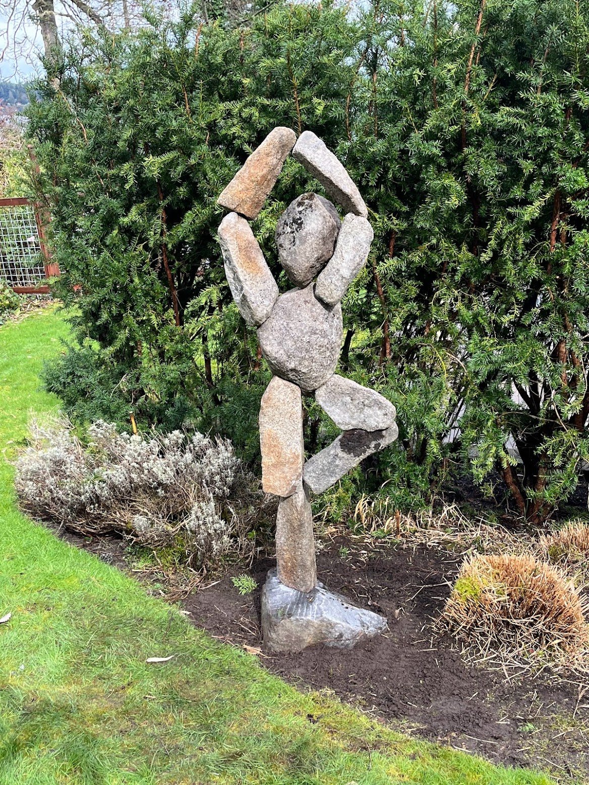 A sculpture made of rocks depicts a human figure with one arm raised above its head, holding a large rock. The sculpture stands in a garden with green bushes and plants in the background.