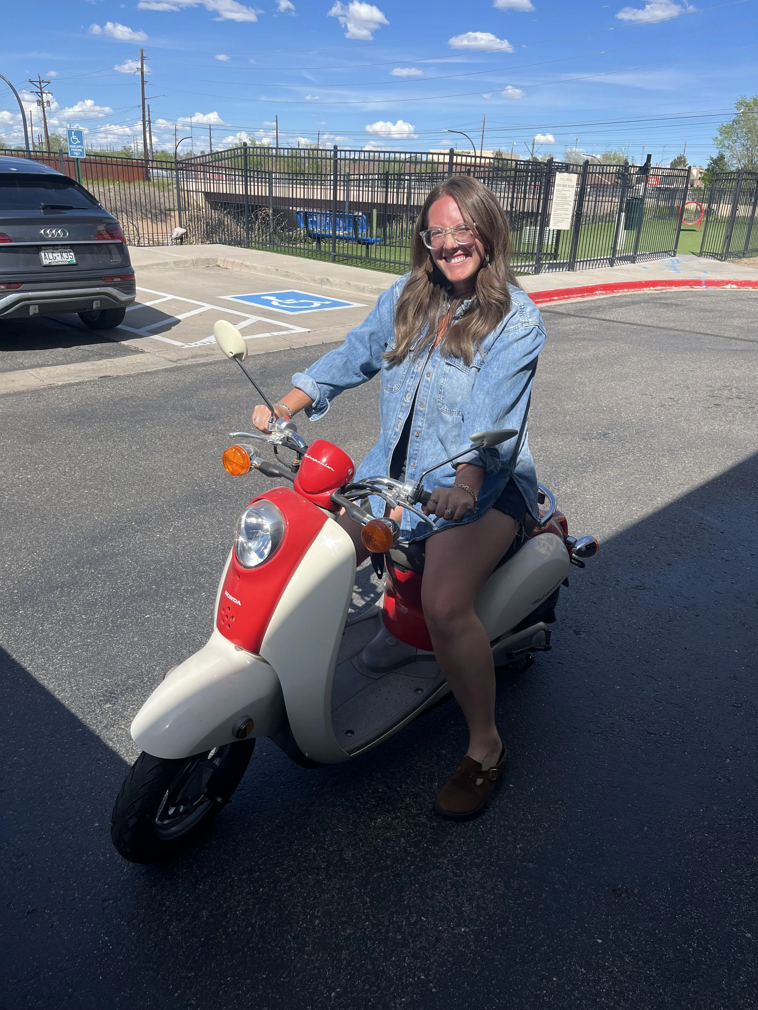 Woman with long hair, glasses, wearing denim jacket and shorts, riding a red and beige scooter on asphalt in a parking lot with cars and a blue handicap parking sign.