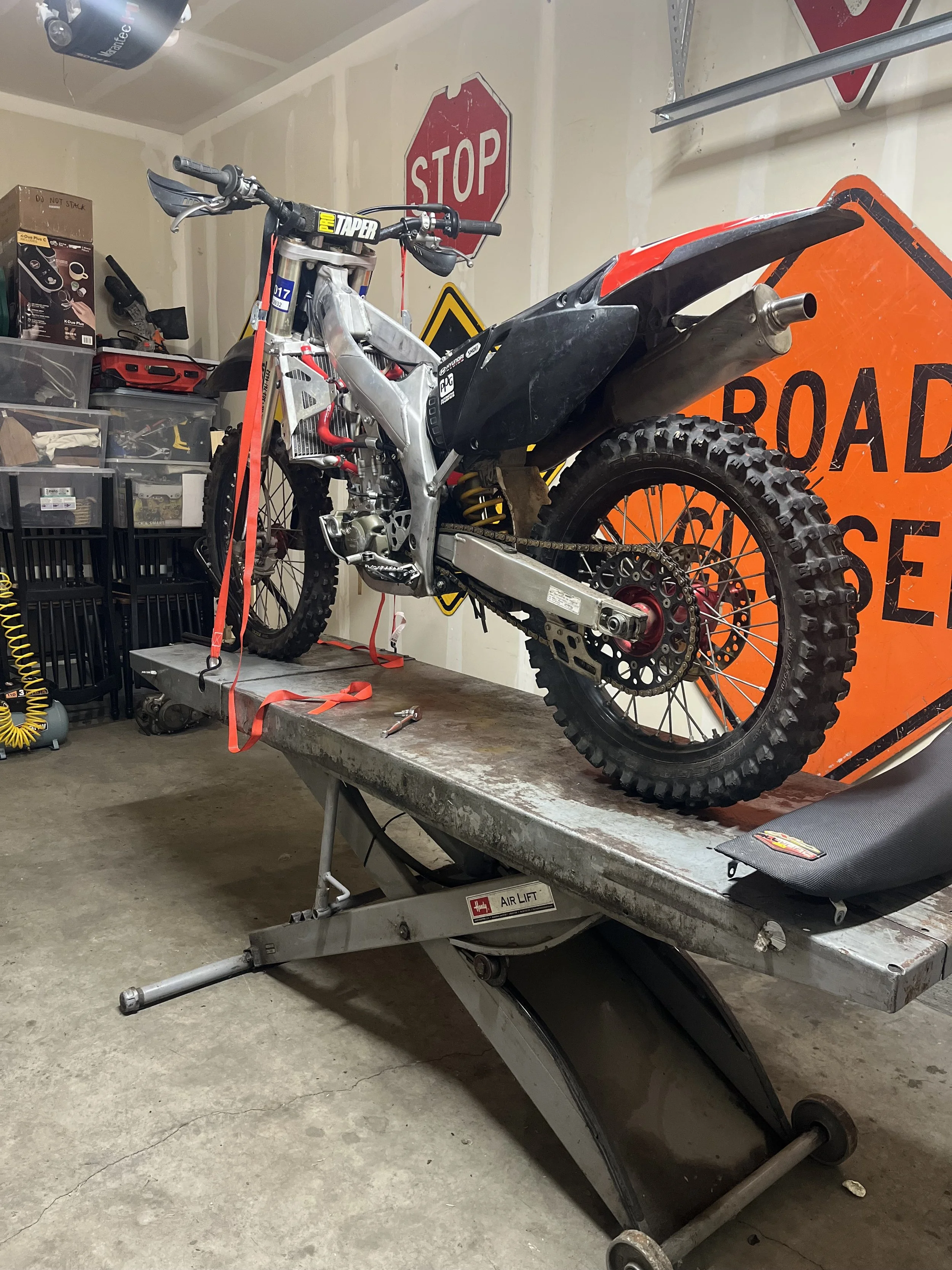 A dirt bike on a work table in a garage with road signs on the wall, including a 'STOP' and an 'Orange Road Work Ahead' sign.