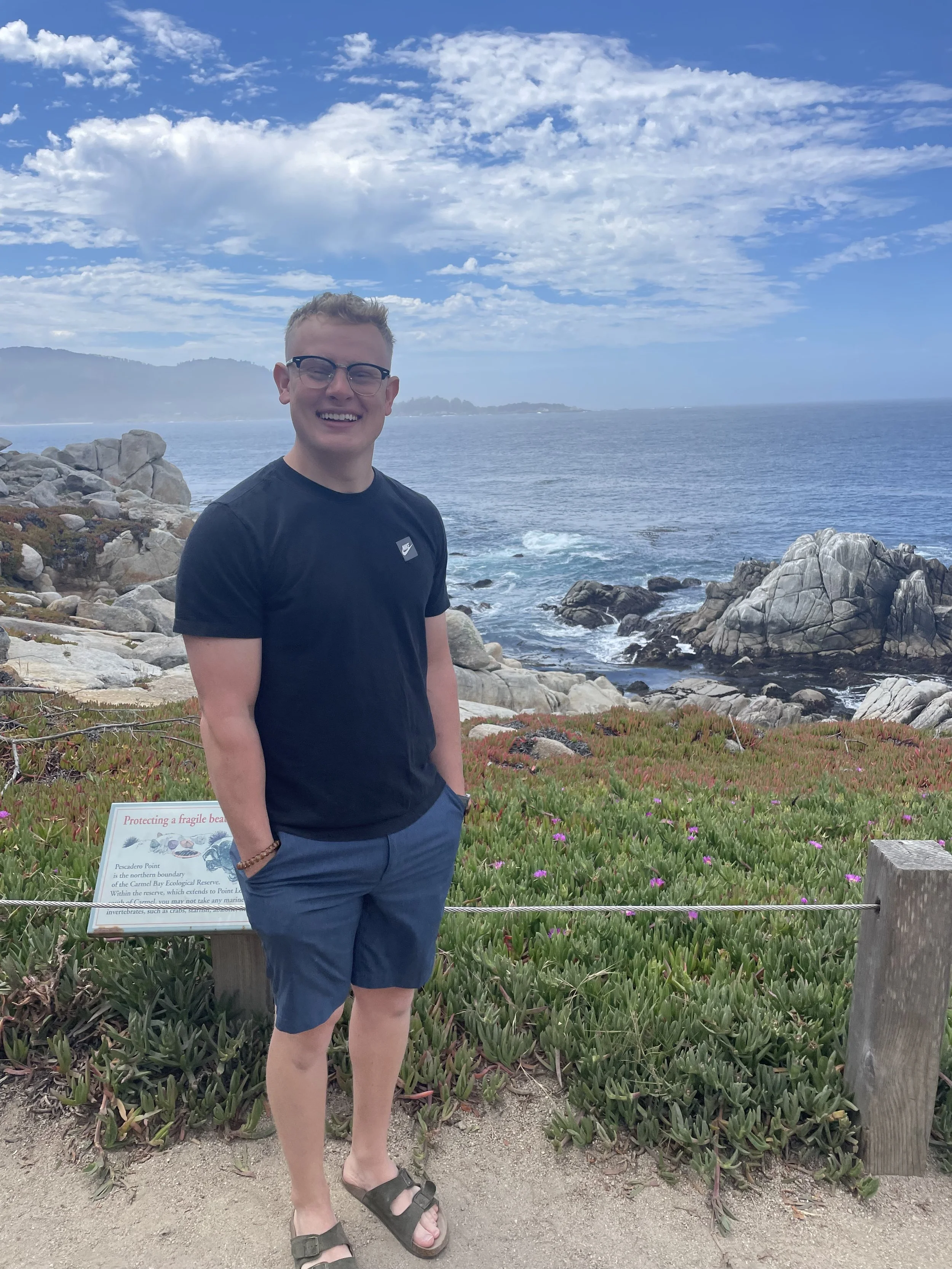 A smiling young man with glasses, wearing a black T-shirt, navy shorts, and sandals, standing on a rocky coast with ocean waves, rocks, and a partly cloudy sky in the background.
