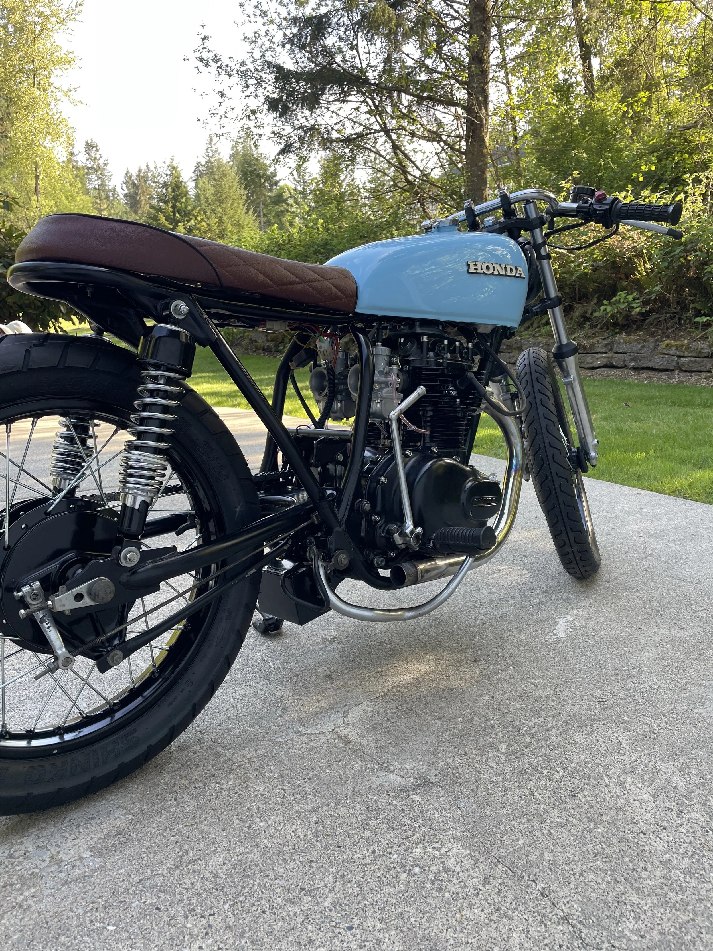 A vintage Honda motorcycle with a light blue gas tank, brown seat, black frame, and black wheels, parked on a concrete driveway surrounded by greenery.