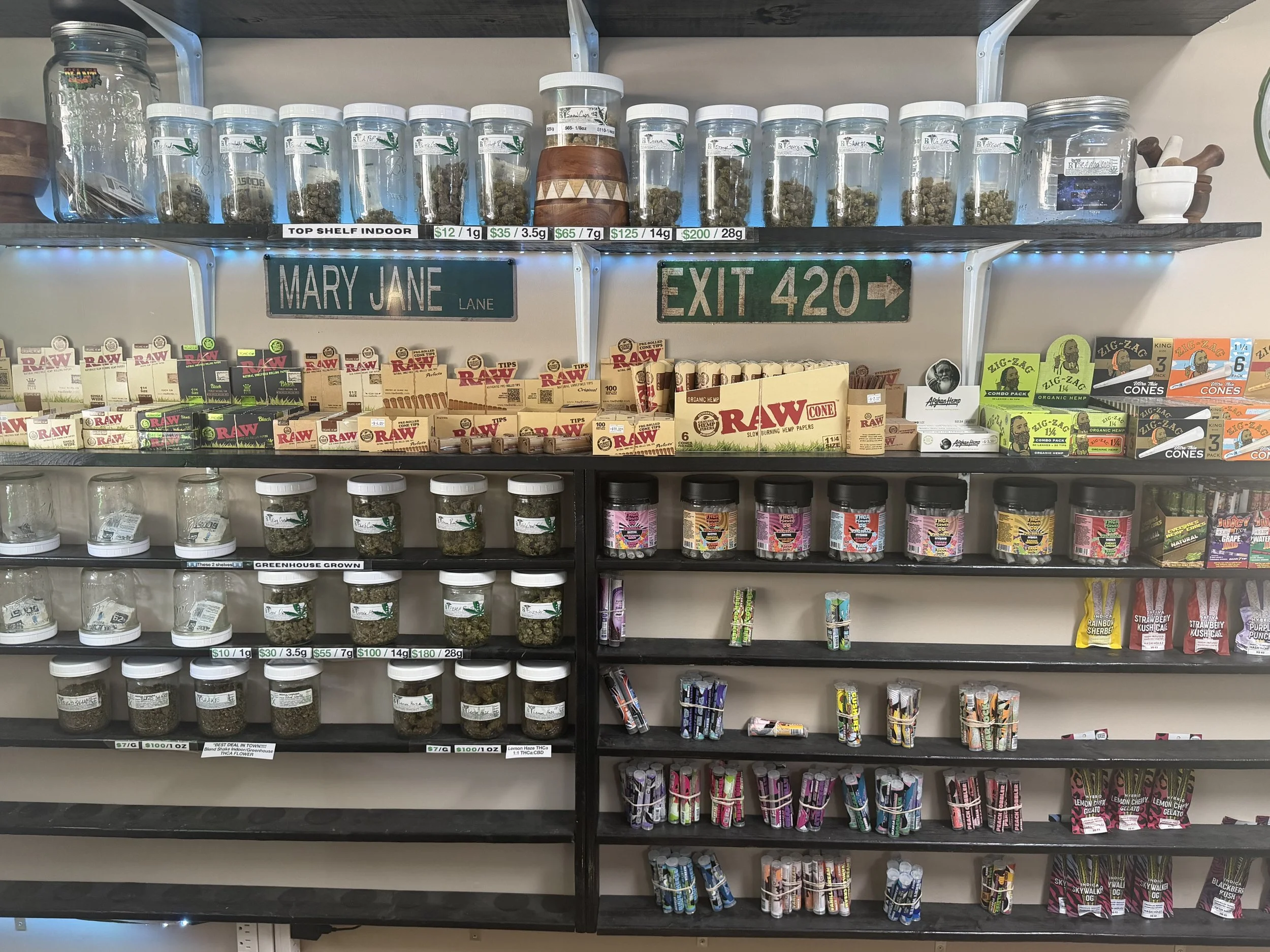 Shelf stocked with jars and packages of cannabis and accessories, with brand labels including RAW and Apple Hemp, along with snack items and a sign that reads "EXIT 420."