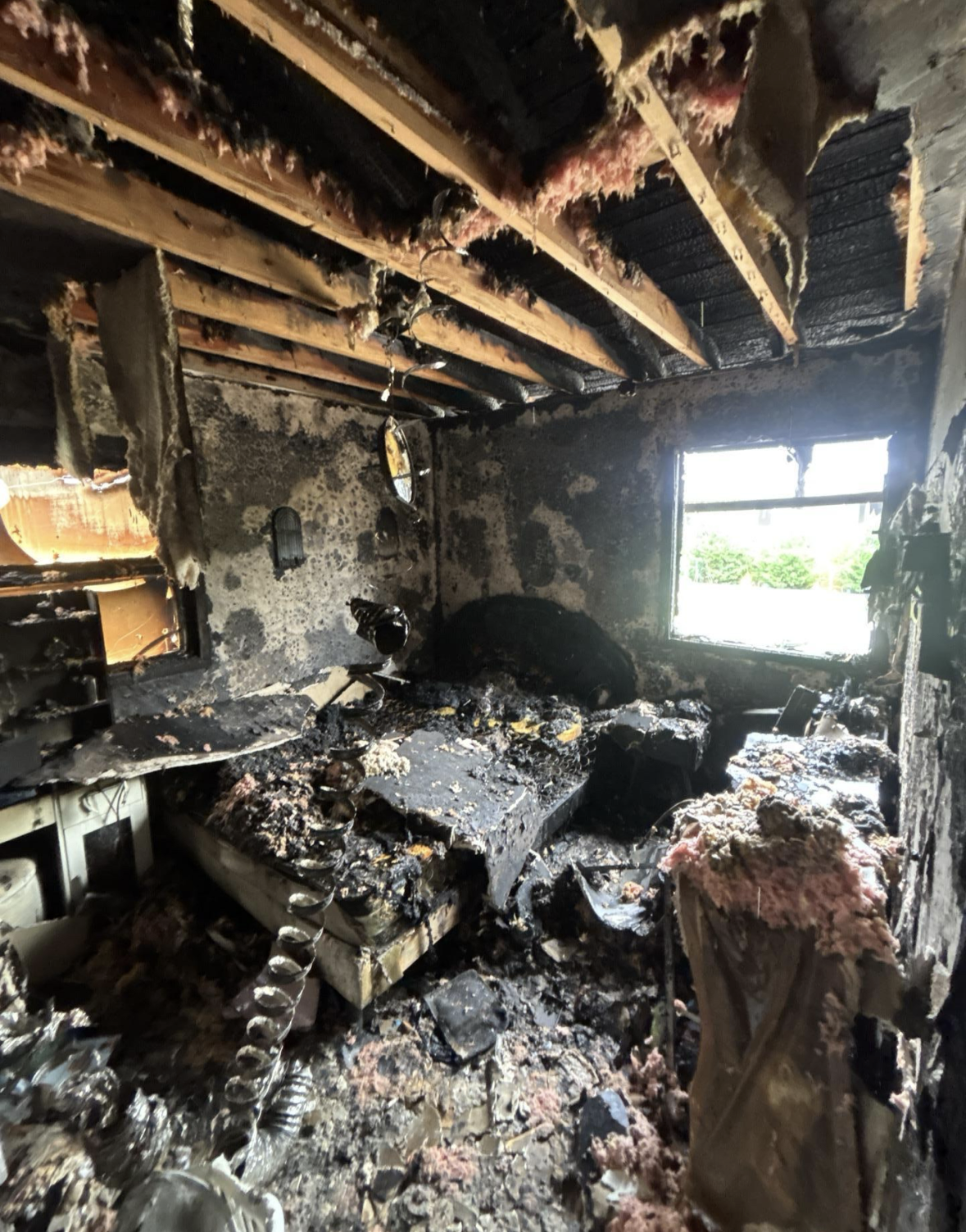 A room severely damaged by fire, with blackened walls, burned furniture, and debris on the floor. The ceiling has exposed wooden beams, some with insulation hanging down, and a window shows daylight outside.