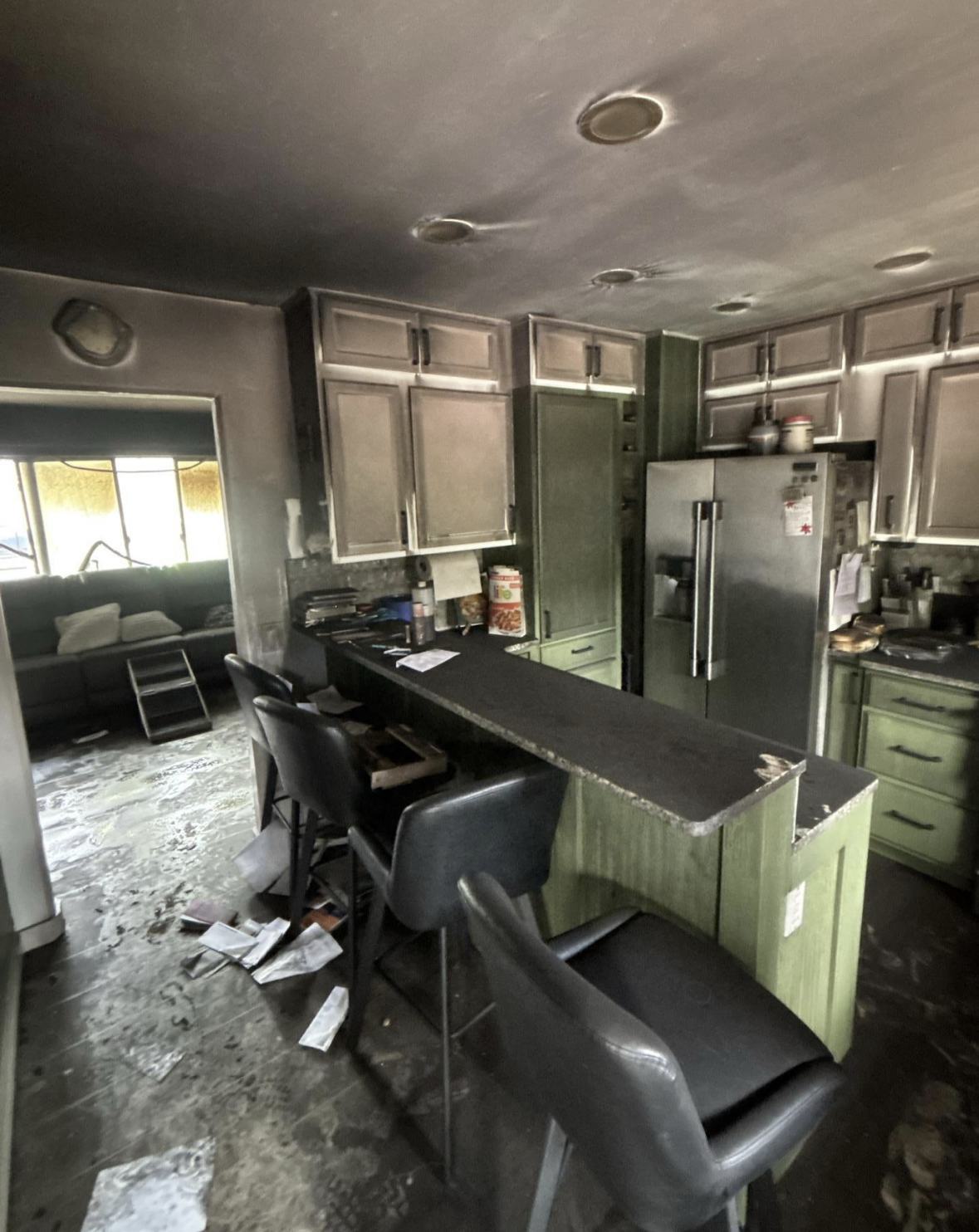 A cluttered kitchen with green cabinets, a black countertop, and black chairs, adjacent to a living room with a sofa, with papers and debris on the floor.