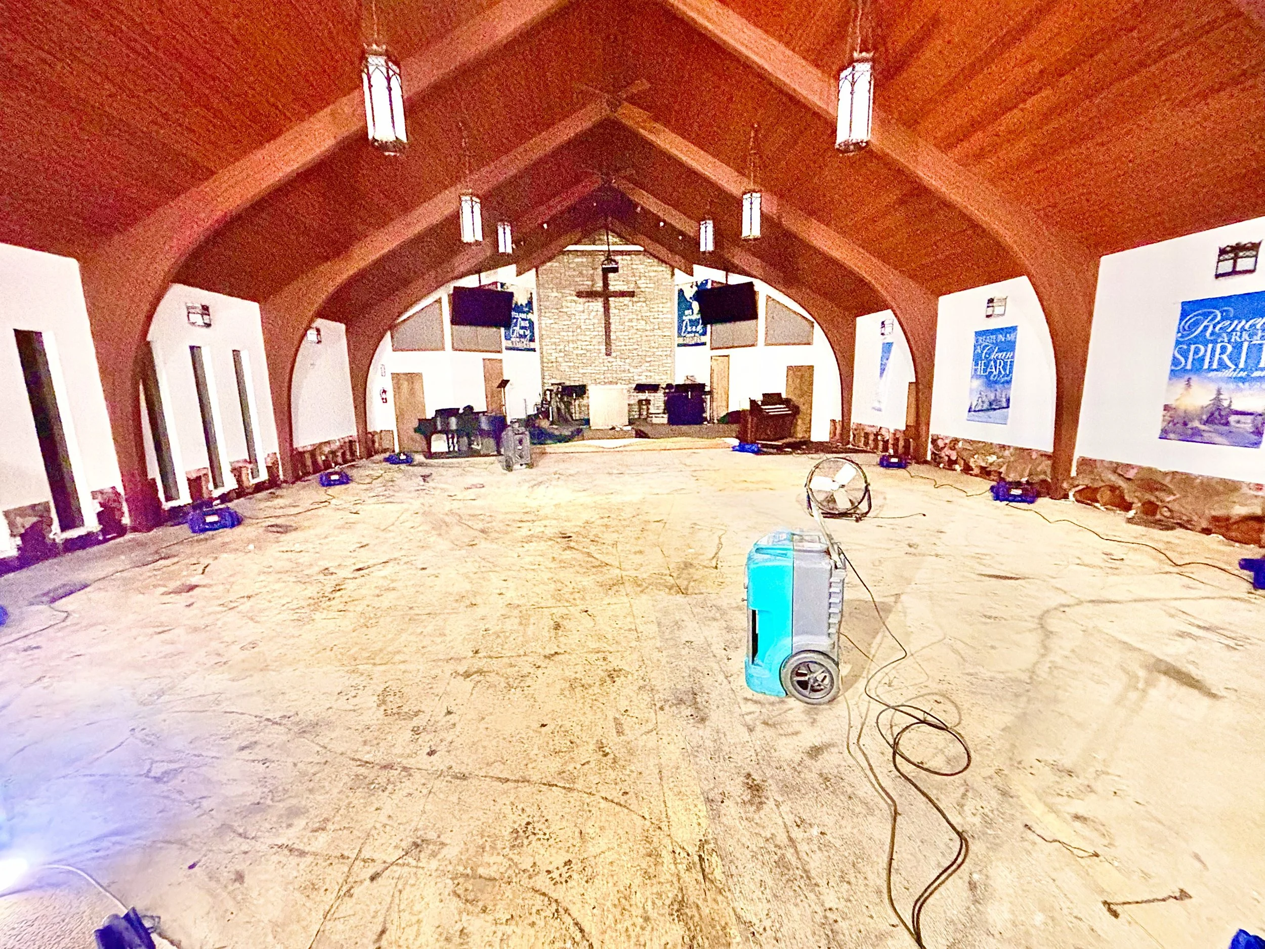 Interior of a church with a wooden vaulted ceiling, a cross at the front, and empty pews along the sides. There are large blue posters on the walls, a stage with musical instruments, and some equipment and cables on the floor.