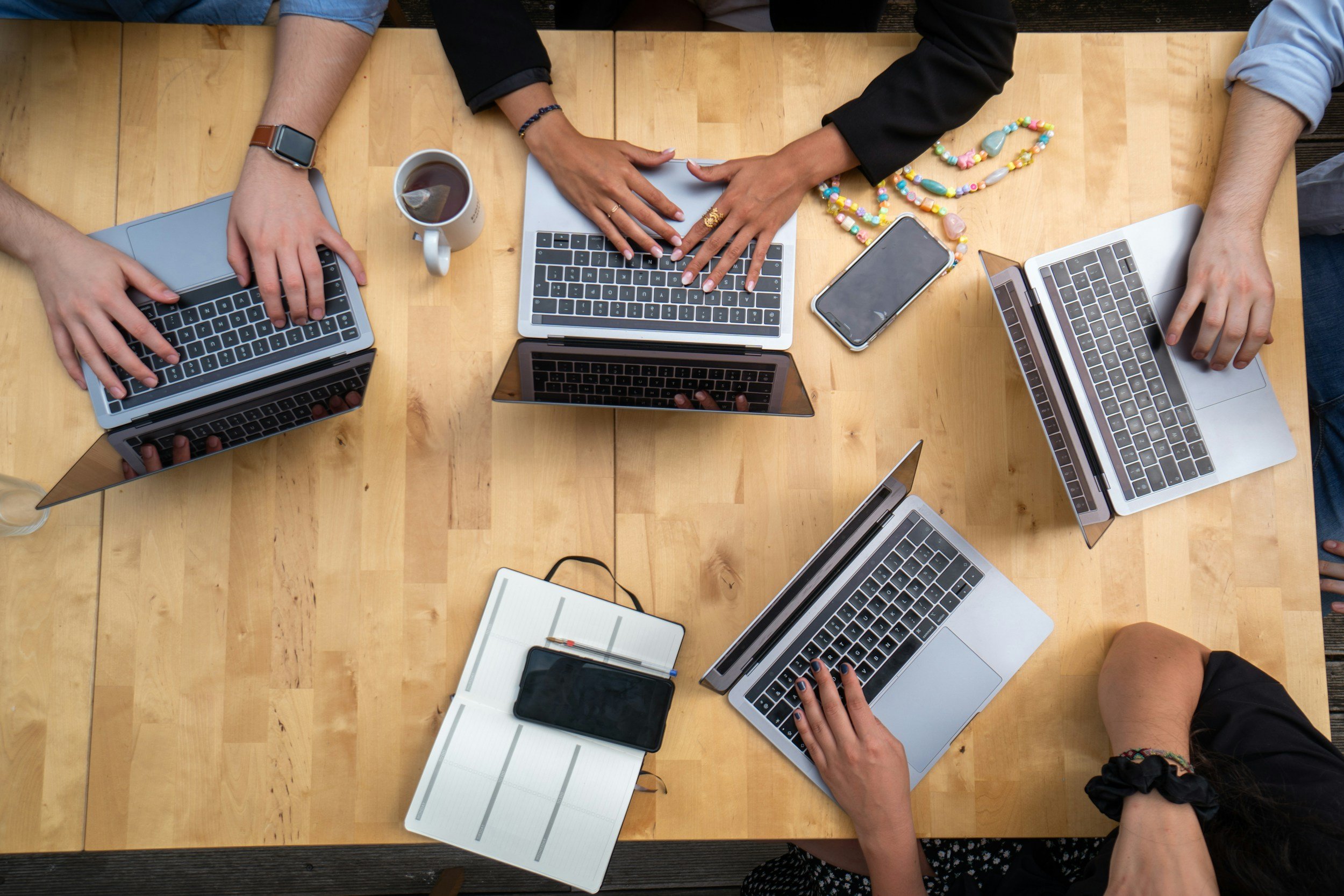 Top-down view of a wooden table with four people using laptops, a coffee mug, a smartphone, a planner with a pen, and colorful necklaces around the table.