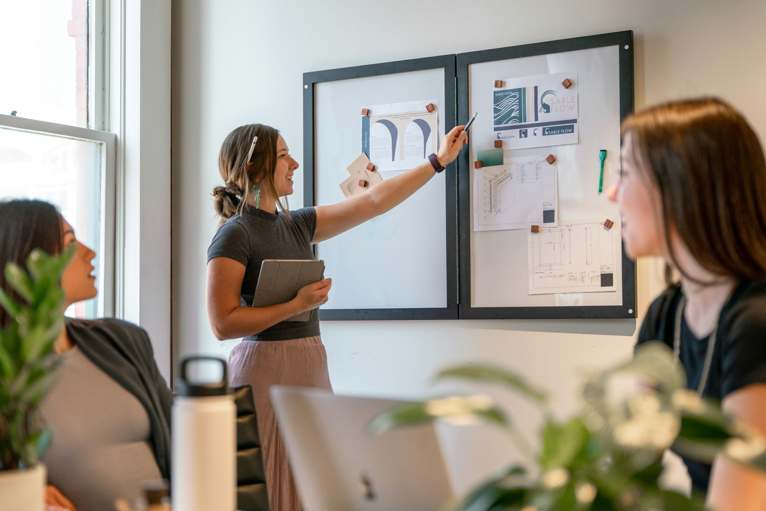 A woman presents to a group at a whiteboard with diagrams and charts in a meeting room, while three women listen attentively.