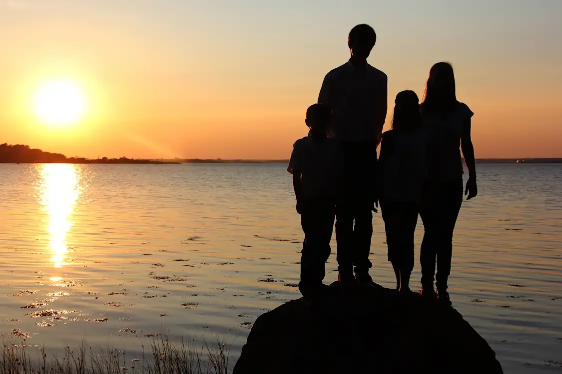 Silhouetted family of four standing on a rock by a lake at sunset.