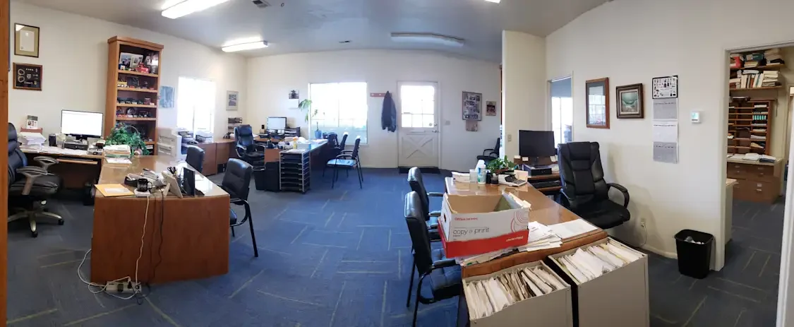 Office room with multiple desks, computers, chairs, and filing cabinets, with papers and office supplies on the desks.