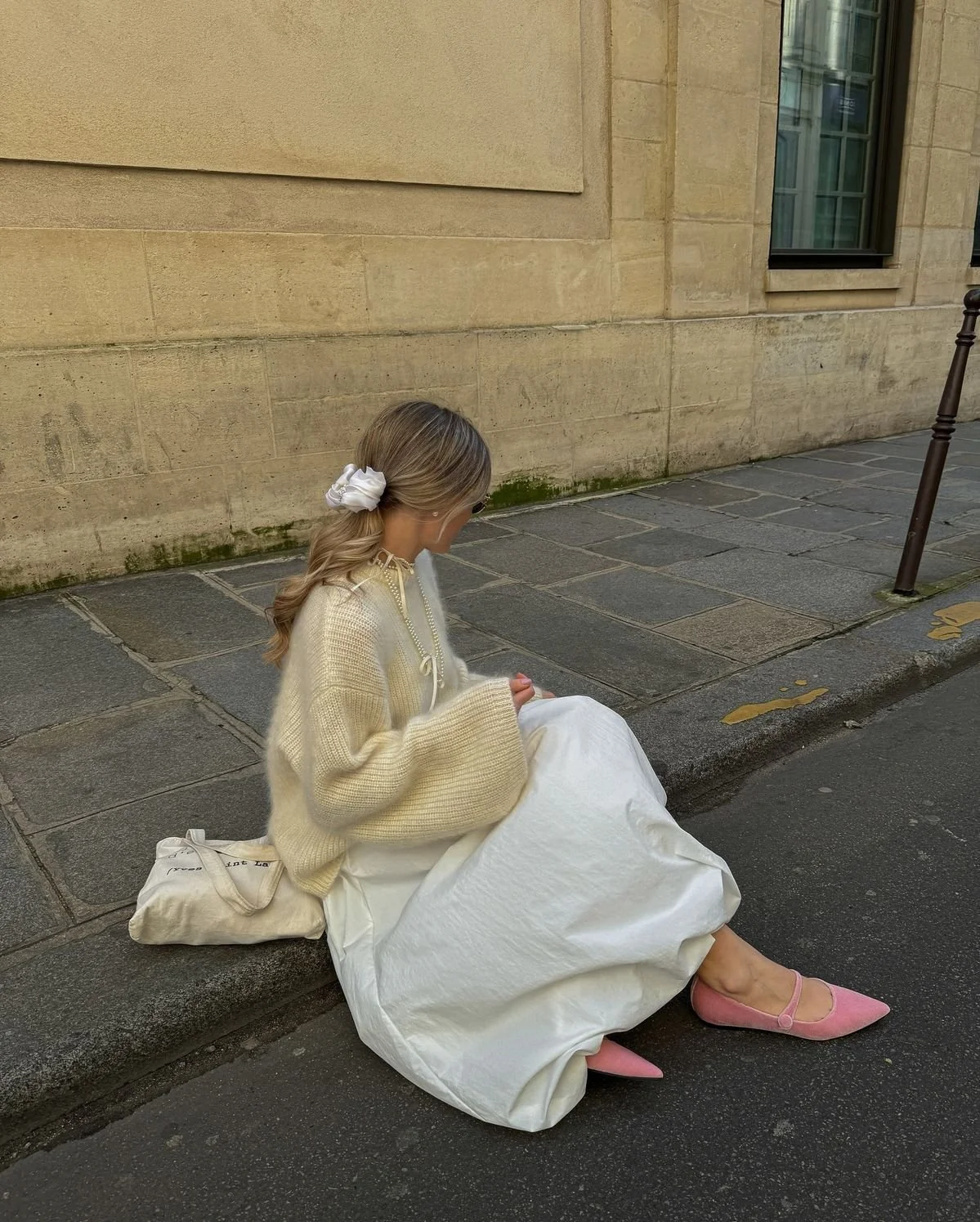 A woman in a cream-colored sweater and long white skirt sitting on a sidewalk with her legs extended, wearing pink pointed-toe flats, with a cream bag beside her, against a beige brick wall.